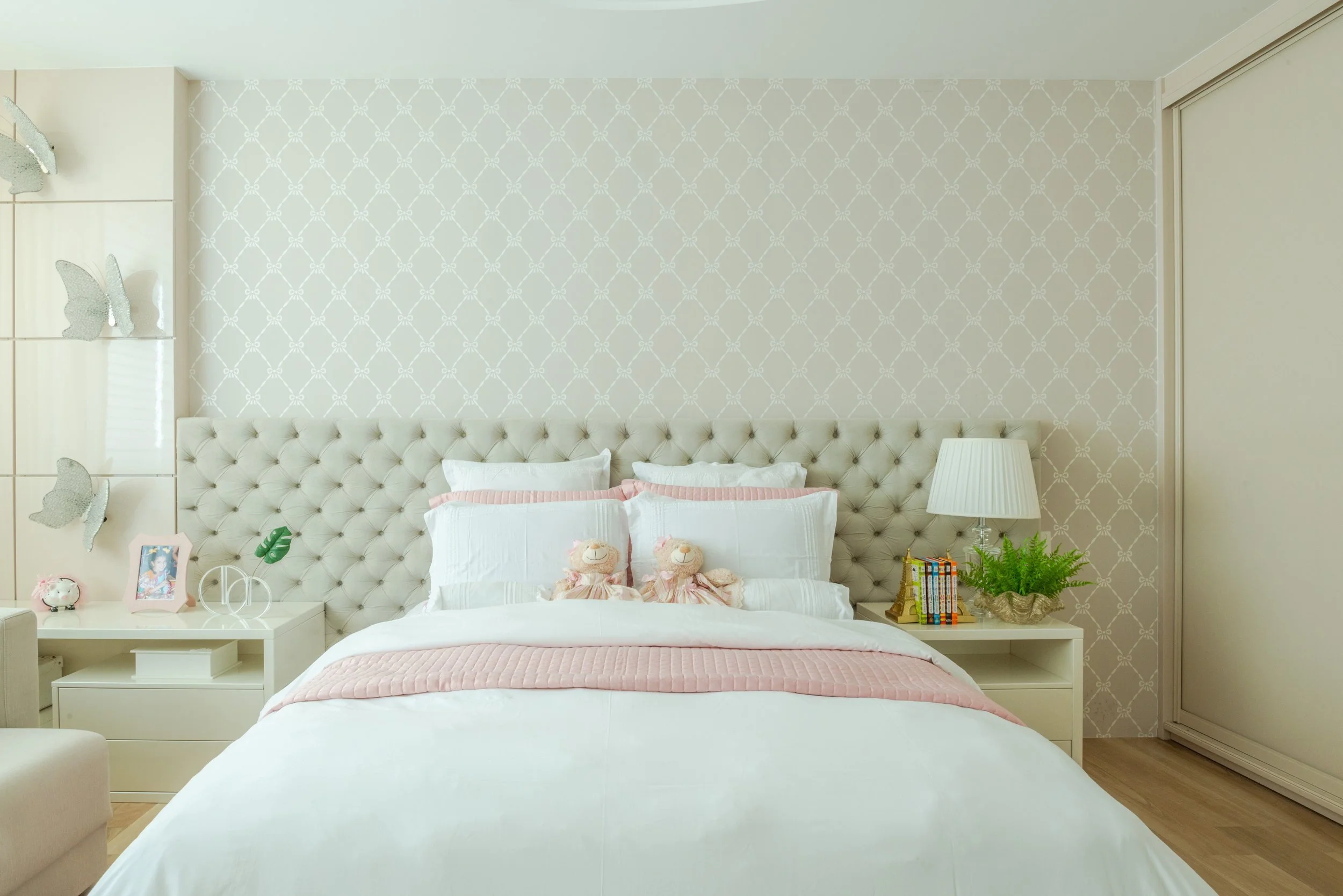 Bedroom with a tufted white headboard, white bedding with pink accents, plush toys, a bedside lamp, and decorative items.