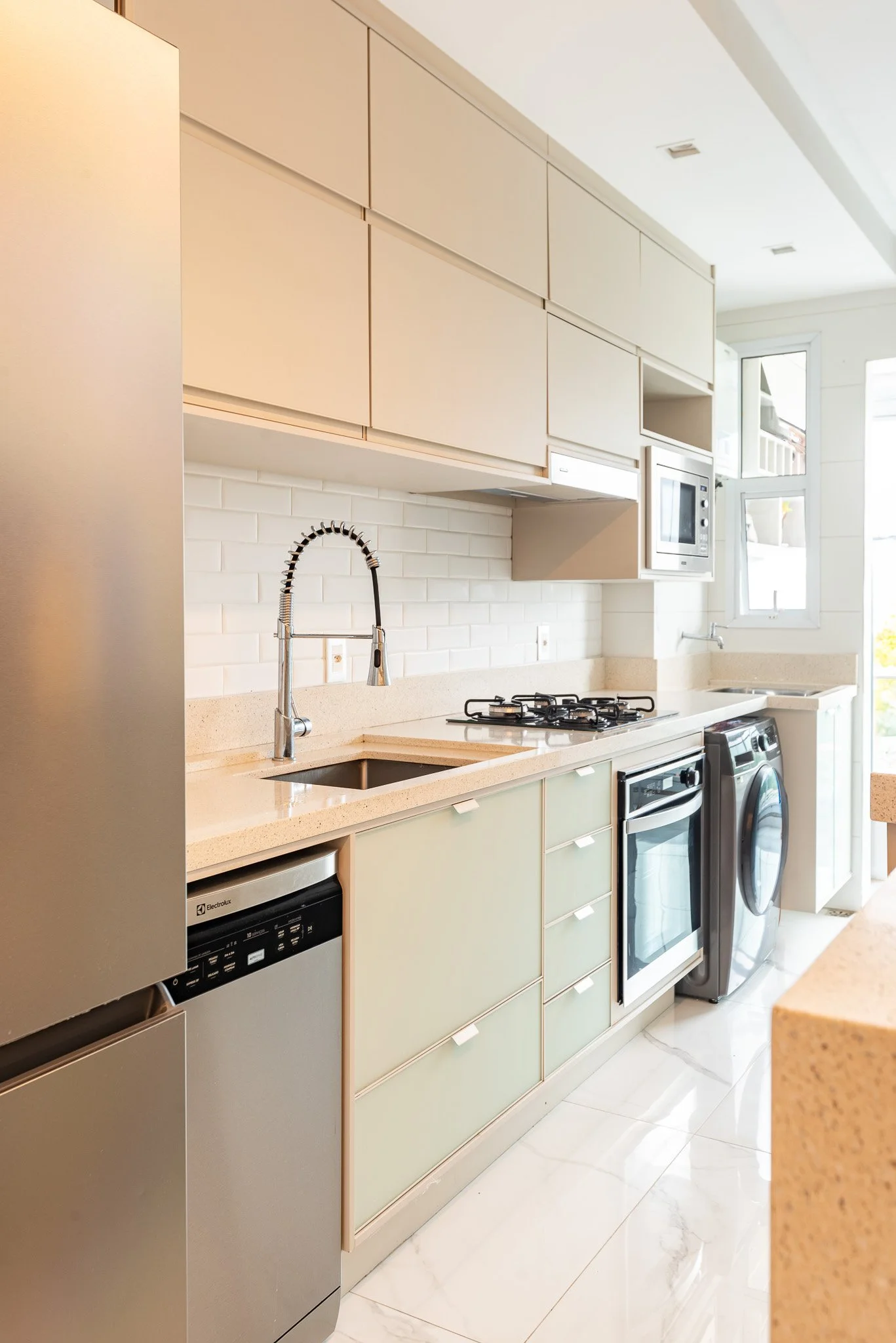 Modern kitchen with beige cabinets, white brick backsplash, beige countertops, a stainless steel sink with a flexible spray faucet, dishwasher, gas stove, microwave, and washing machine. Large windows allow natural light.