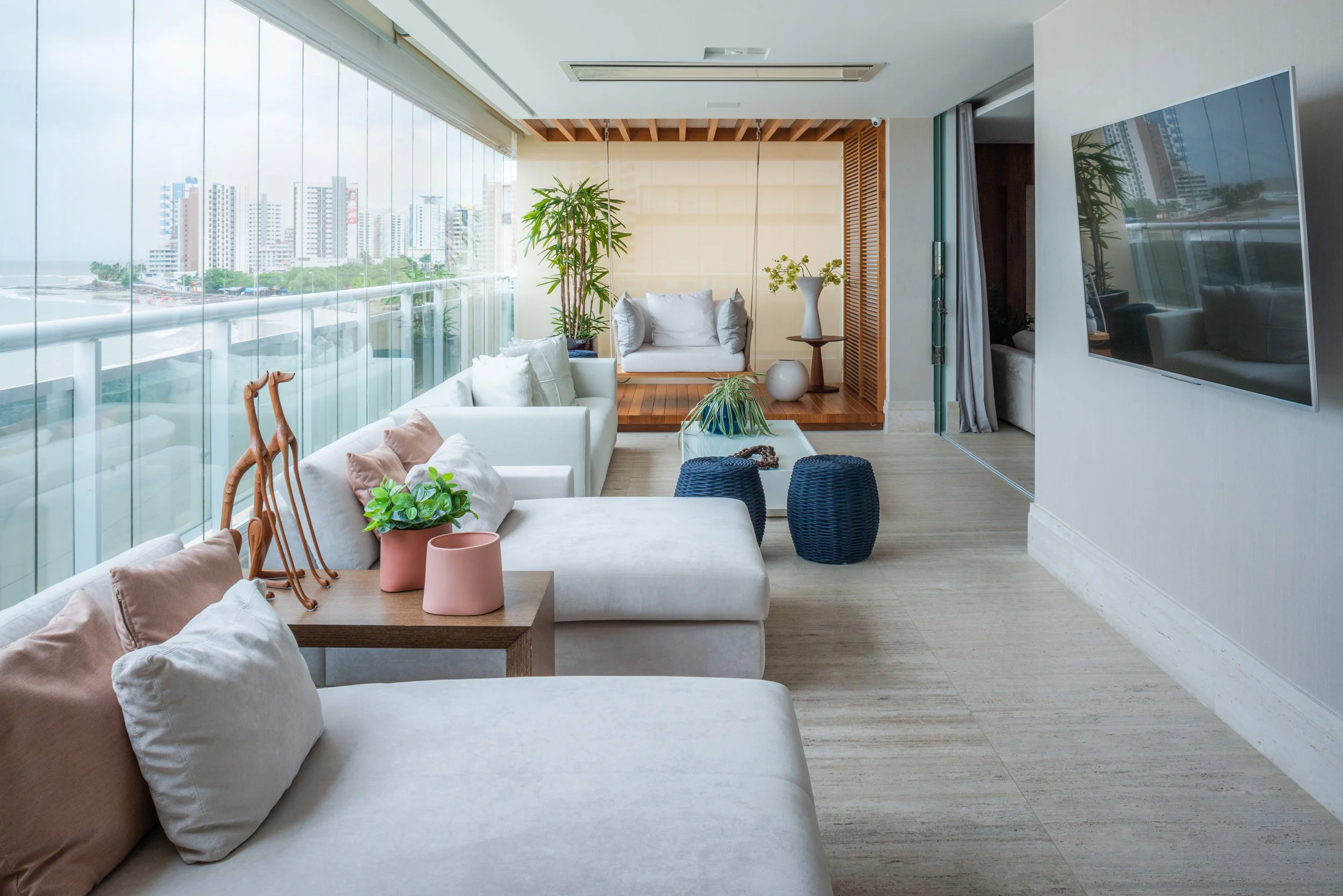Modern living room with large windows, white sofas, potted plants, a wall-mounted TV, and cityscape view.