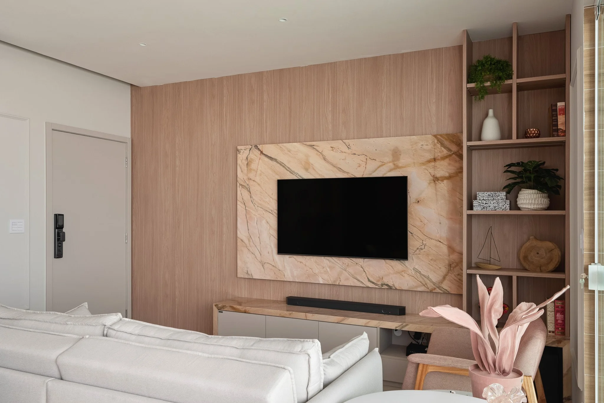 Living room with a mounted flat-screen TV on a marble accent wall, wooden shelves with decorative items, and a white sofa with a pink cushion and plant in a pink pot.