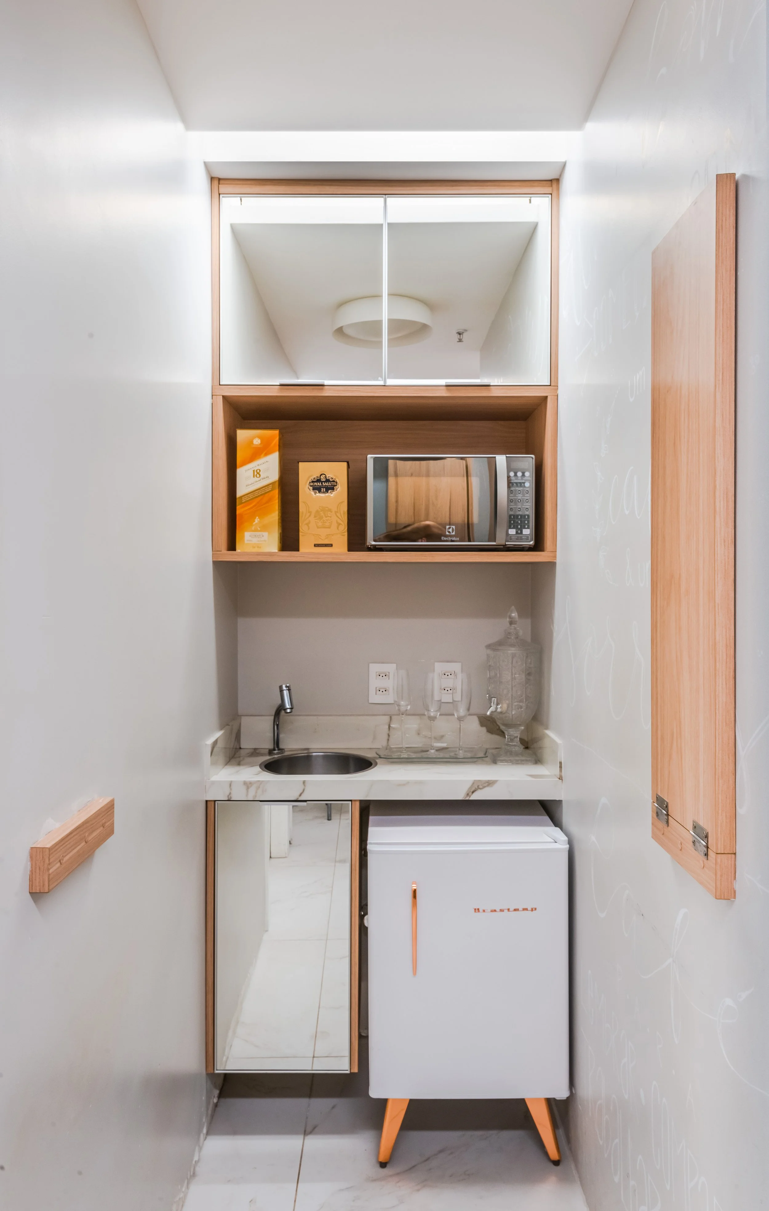 A compact kitchenette with a small sink, mini fridge, microwave, and shelves for tea and coffee supplies, in a narrow space with white walls and marble countertop.