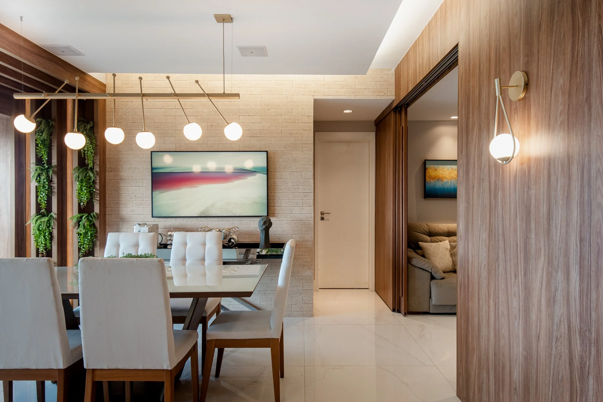 Modern dining room with white upholstered chairs, a glass-top dining table, and a wall-mounted television, featuring wooden and brick interior design elements and hanging globe light fixtures.
