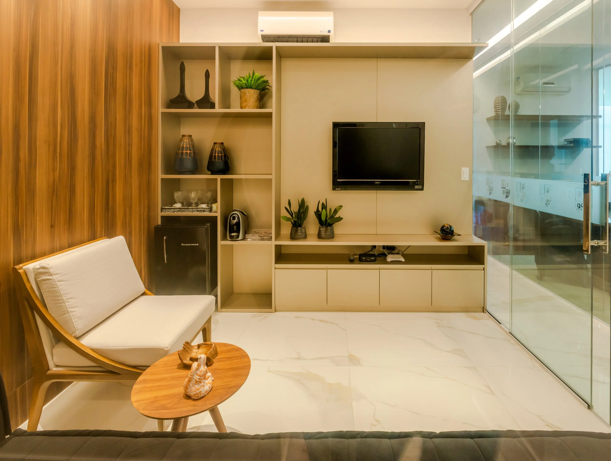 Modern living room with a flat-screen TV, a beige entertainment unit, and a wooden armchair with white cushion. A small wooden coffee table holds decorative items, and glass doors on the right lead to another room.