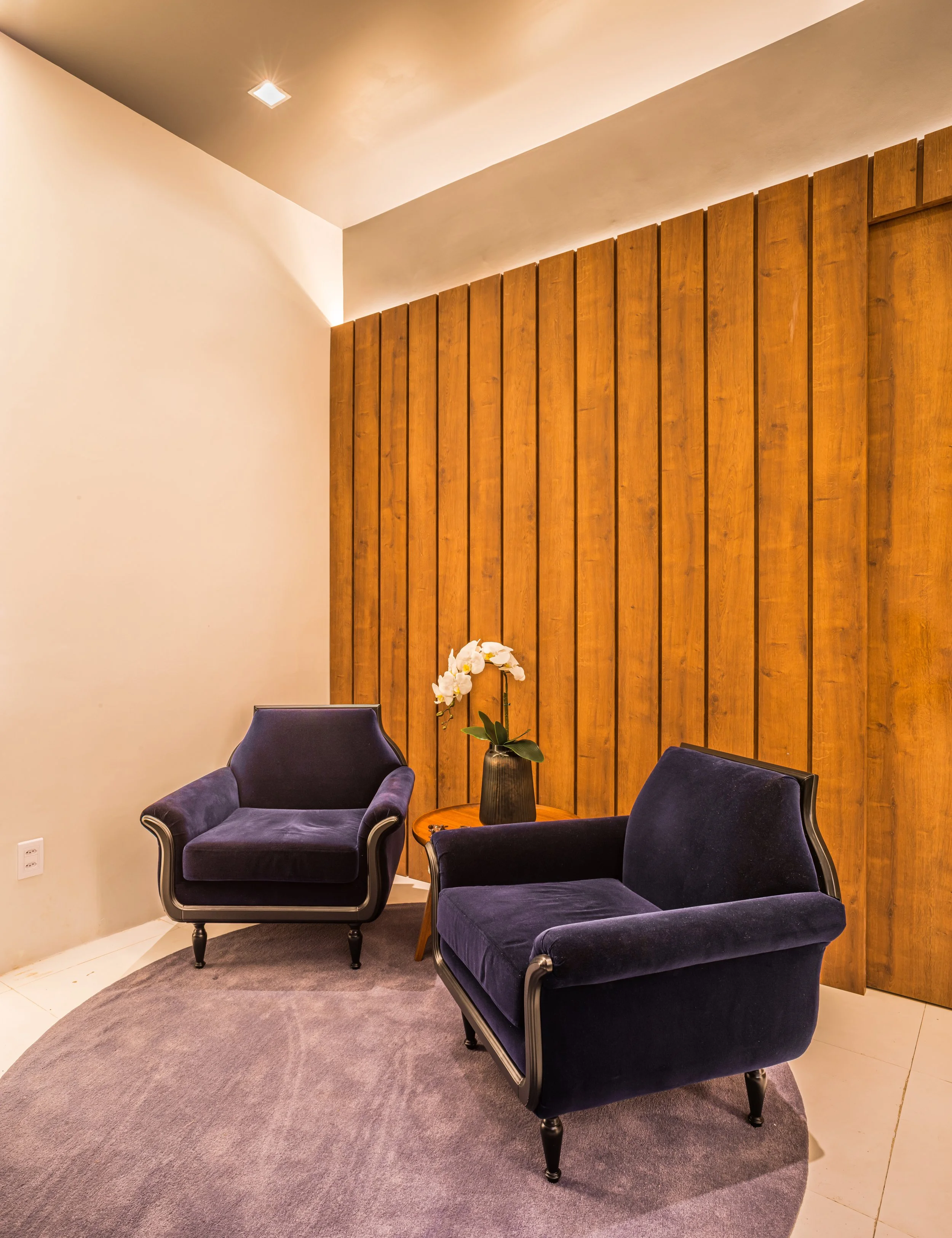 Two dark blue velvet armchairs with black wooden legs, a small round wooden table with a potted white orchid, wooden wall paneling, beige walls, and ceiling with a recessed light.