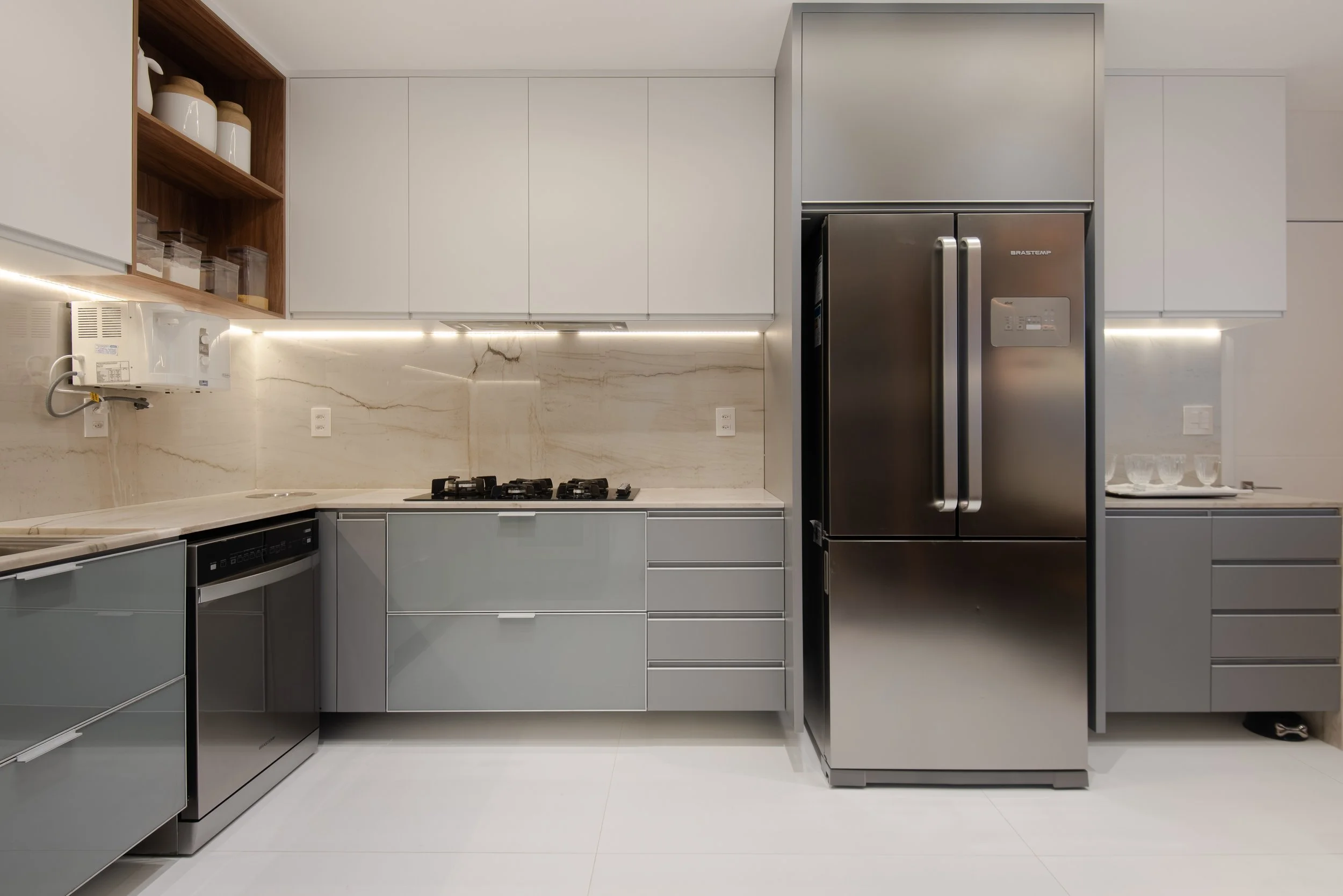 Modern kitchen with gray cabinets, stainless steel refrigerator, built-in oven, marble backsplash, open wooden shelf with jars, and minimalistic decor.