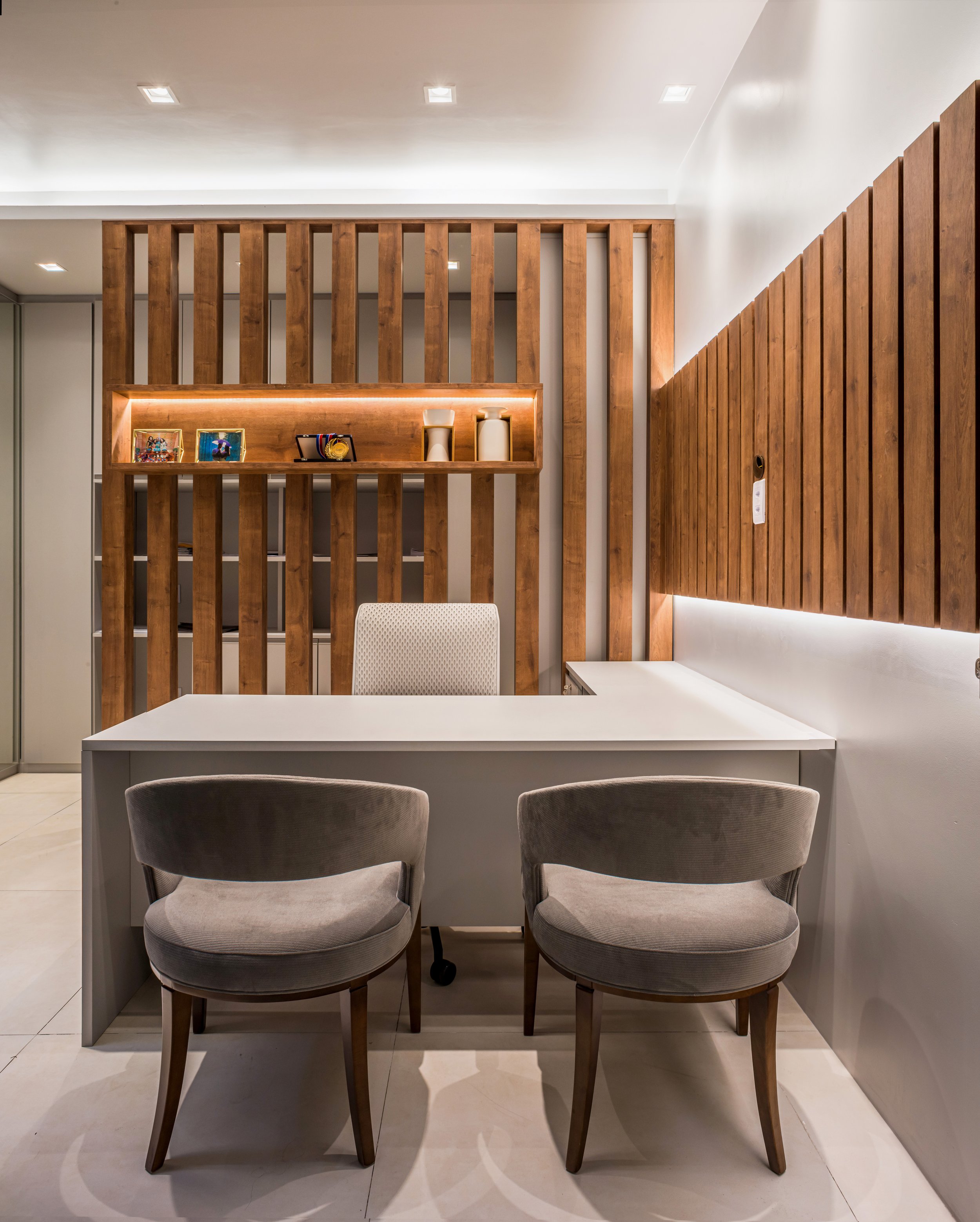 Office reception area with a white desk, two beige chairs, wooden wall paneling, and decorative items on a built-in wooden shelf.