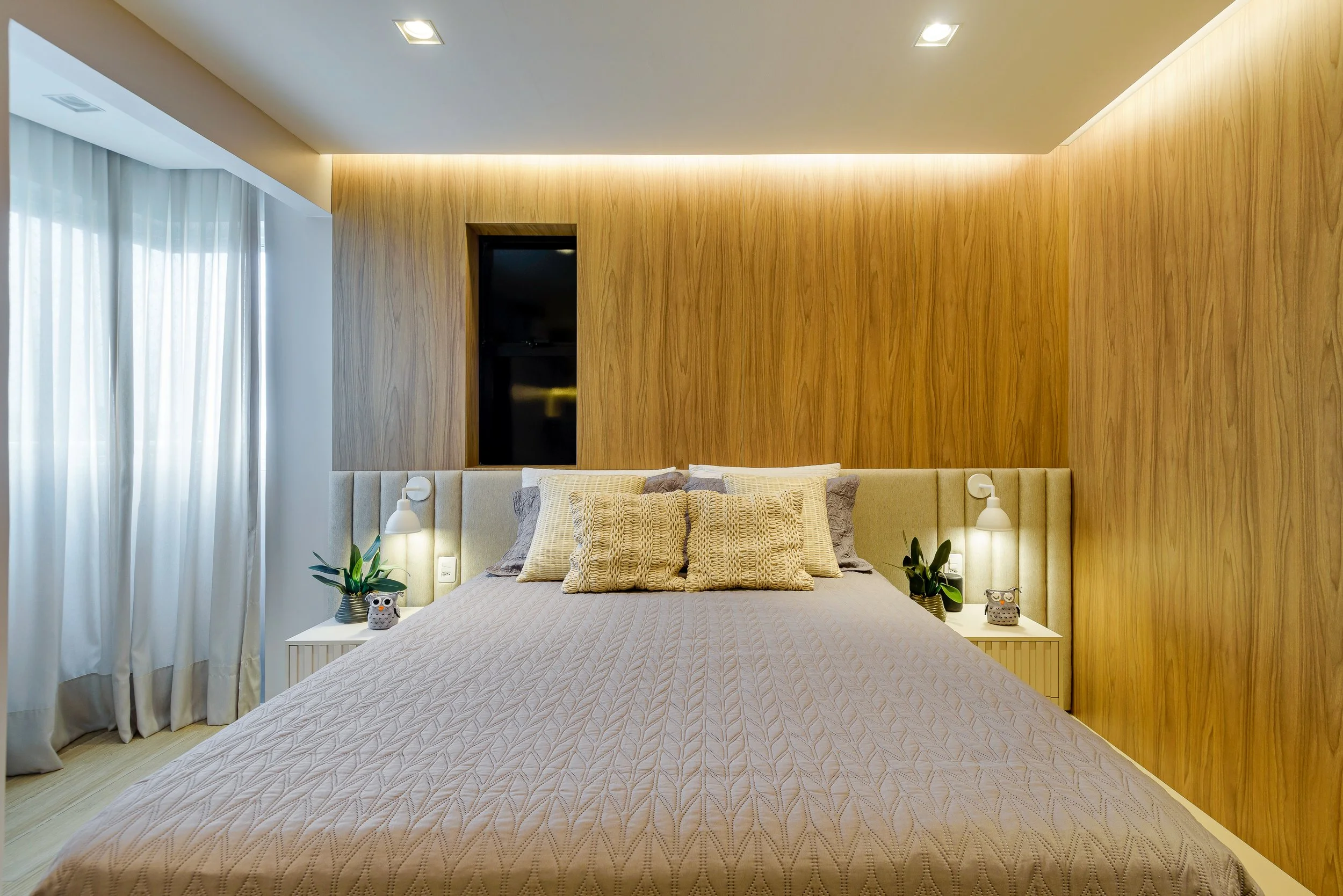 Modern bedroom with beige bed, pillows, wooden accent wall, plant, and lamps.