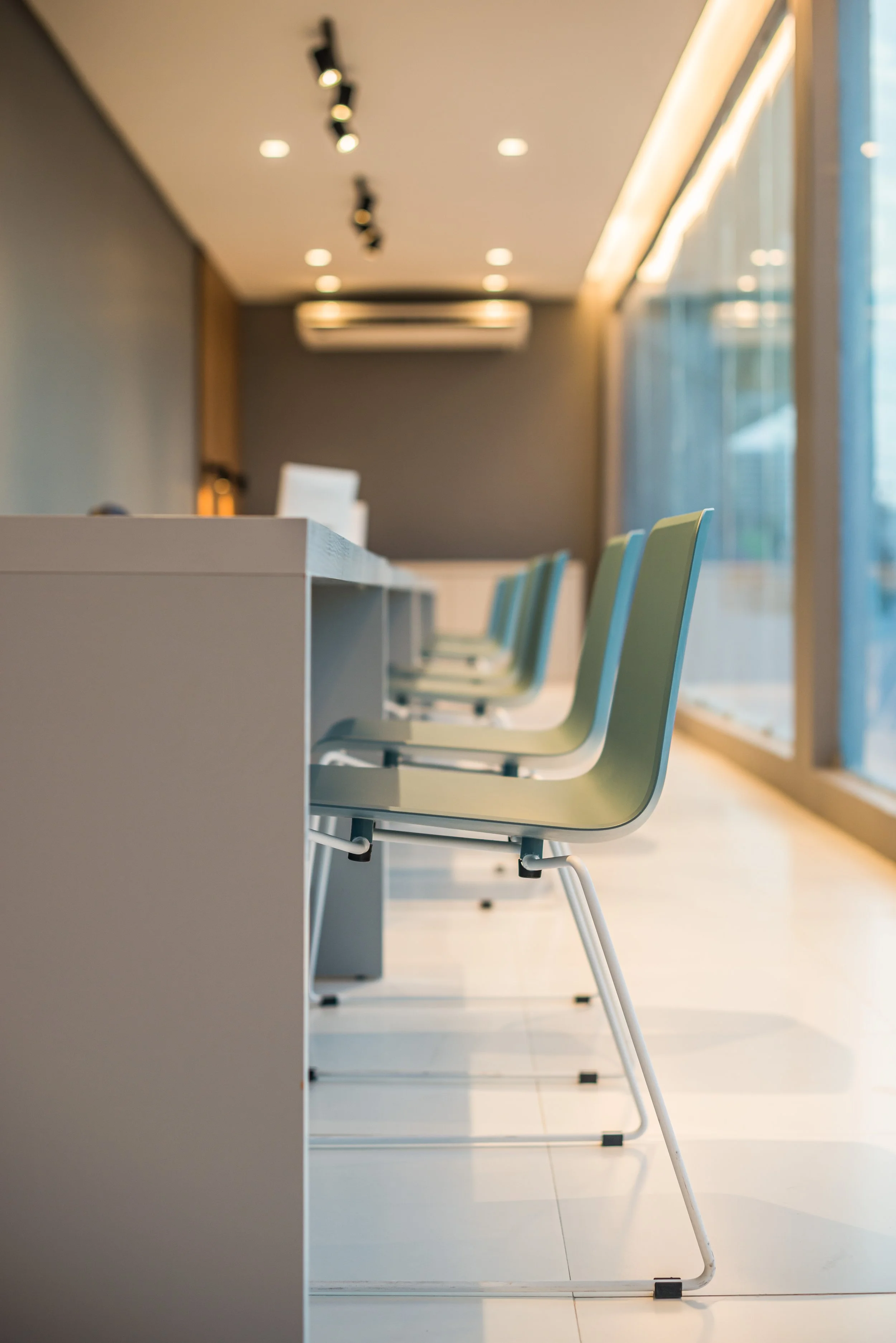 Empty green chairs lined up along a white table in a modern, well-lit conference room with large windows.