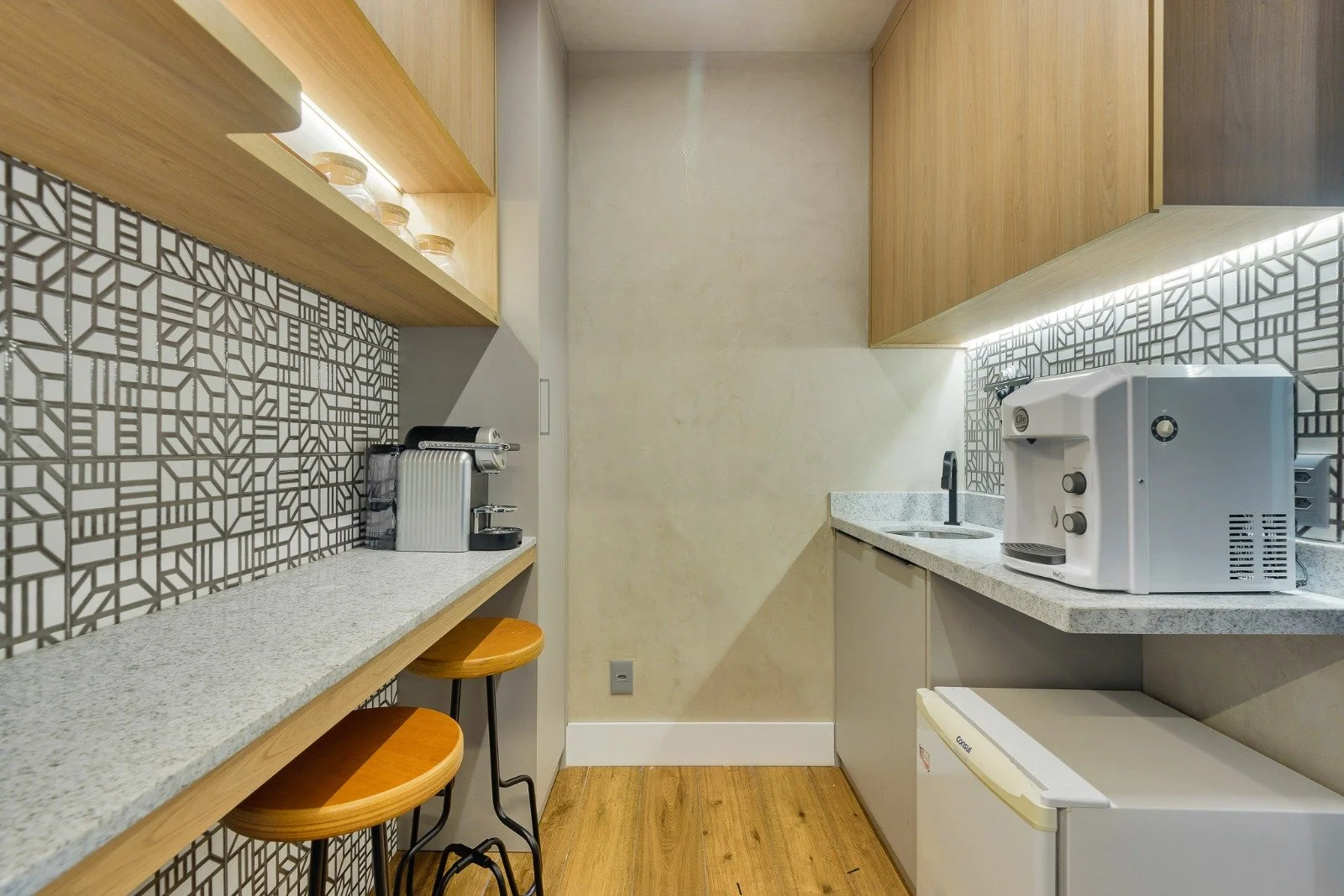 Small modern kitchenette with wooden cabinets, geometric tile backsplash, granite countertops, coffee machine, and mini fridge.