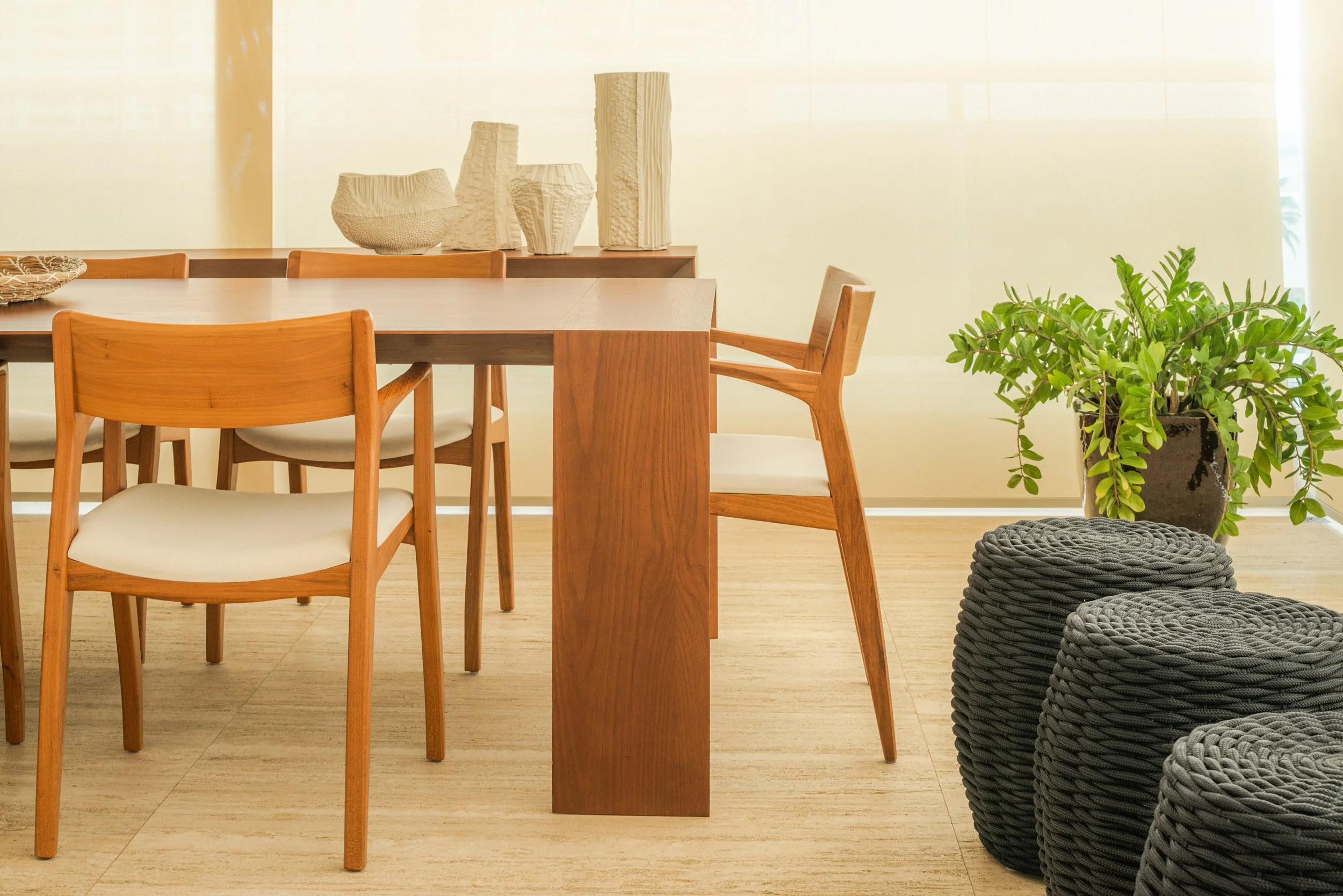 A dining room with a wooden table surrounded by wooden chairs with white cushions, decorative ceramic vases on the table, a large green potted plant, and three round woven gray stools.