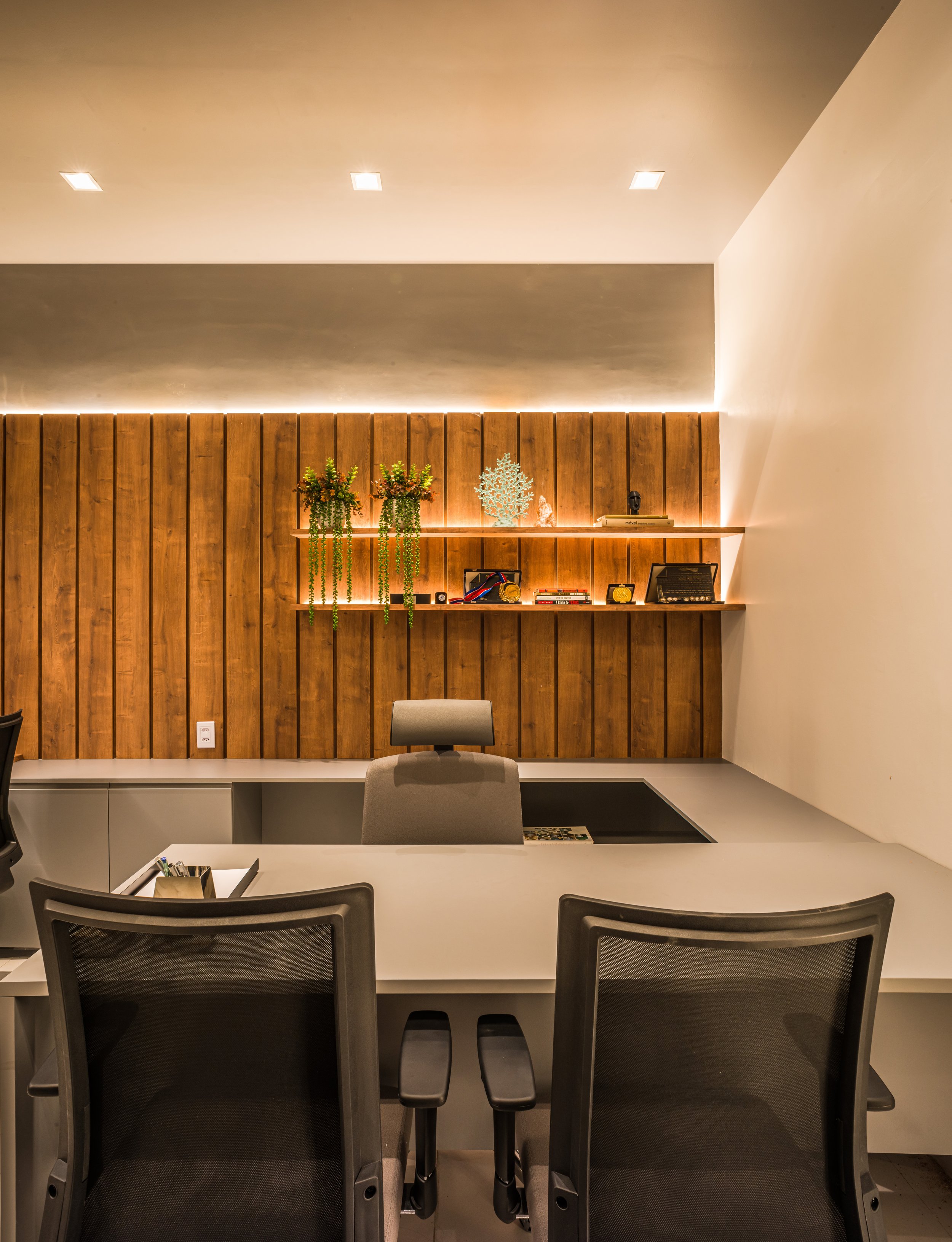 An office workspace with a white desk, two black office chairs, a grey office chair behind the desk, a wooden wall with shelves, plants, and decorative items, under ceiling lights.
