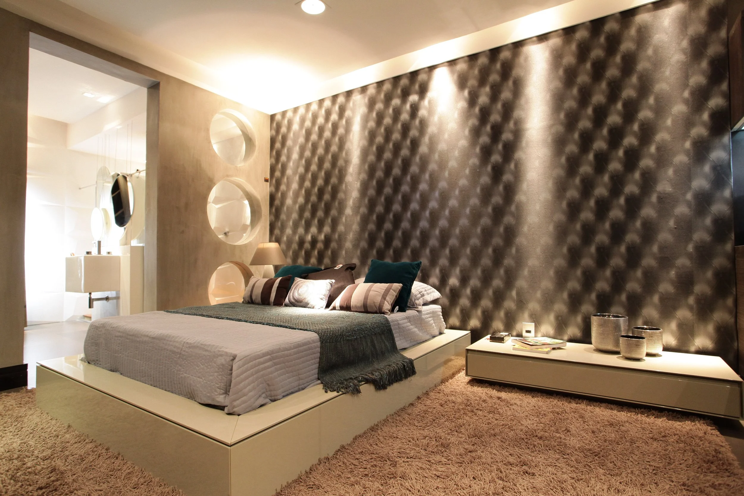 Modern bedroom with a textured dark accent wall, a low platform bed with white bedding and multiple pillows, a bedside lamp, a white dresser with decorative bowls, and a plush beige rug.