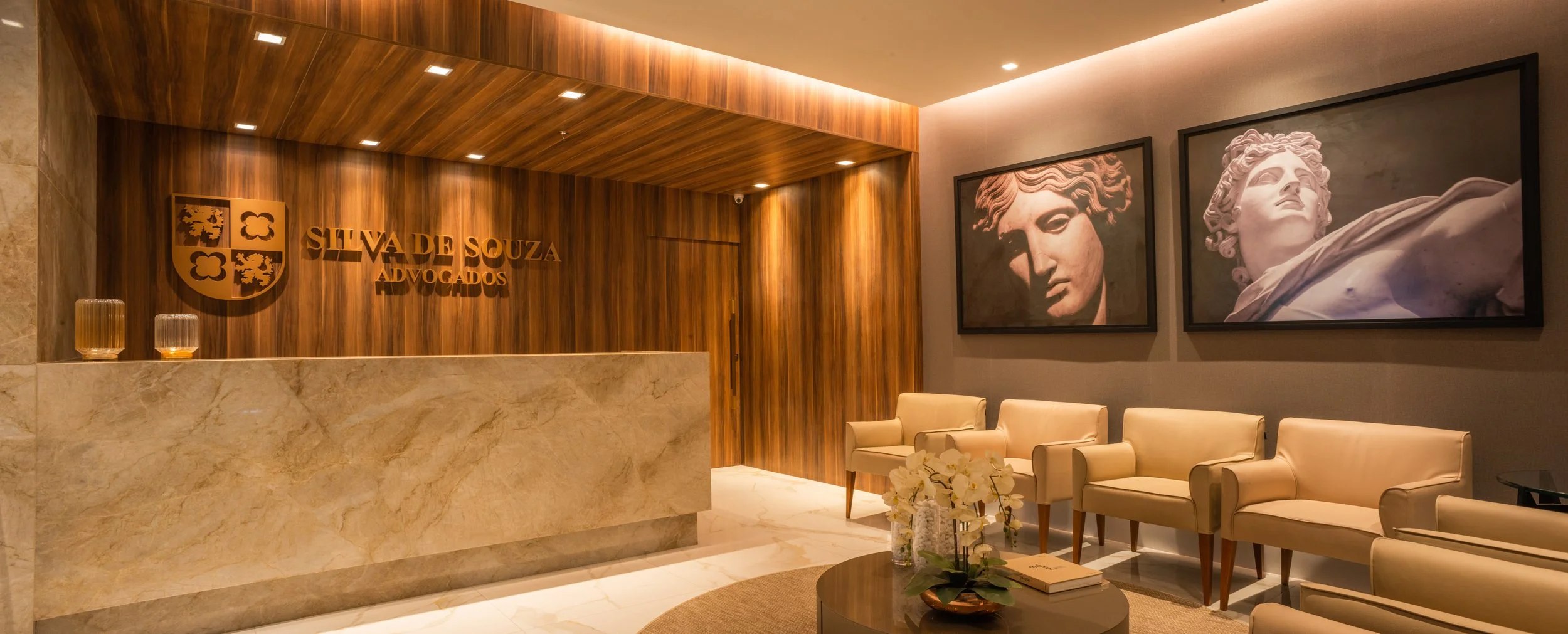 Lobby of Silva de Souza Advocados with a marble reception desk, wooden wall with a coat of arms emblem, and framed classical sculpture portraits on the wall, beige chairs, and a coffee table with flowers and books.