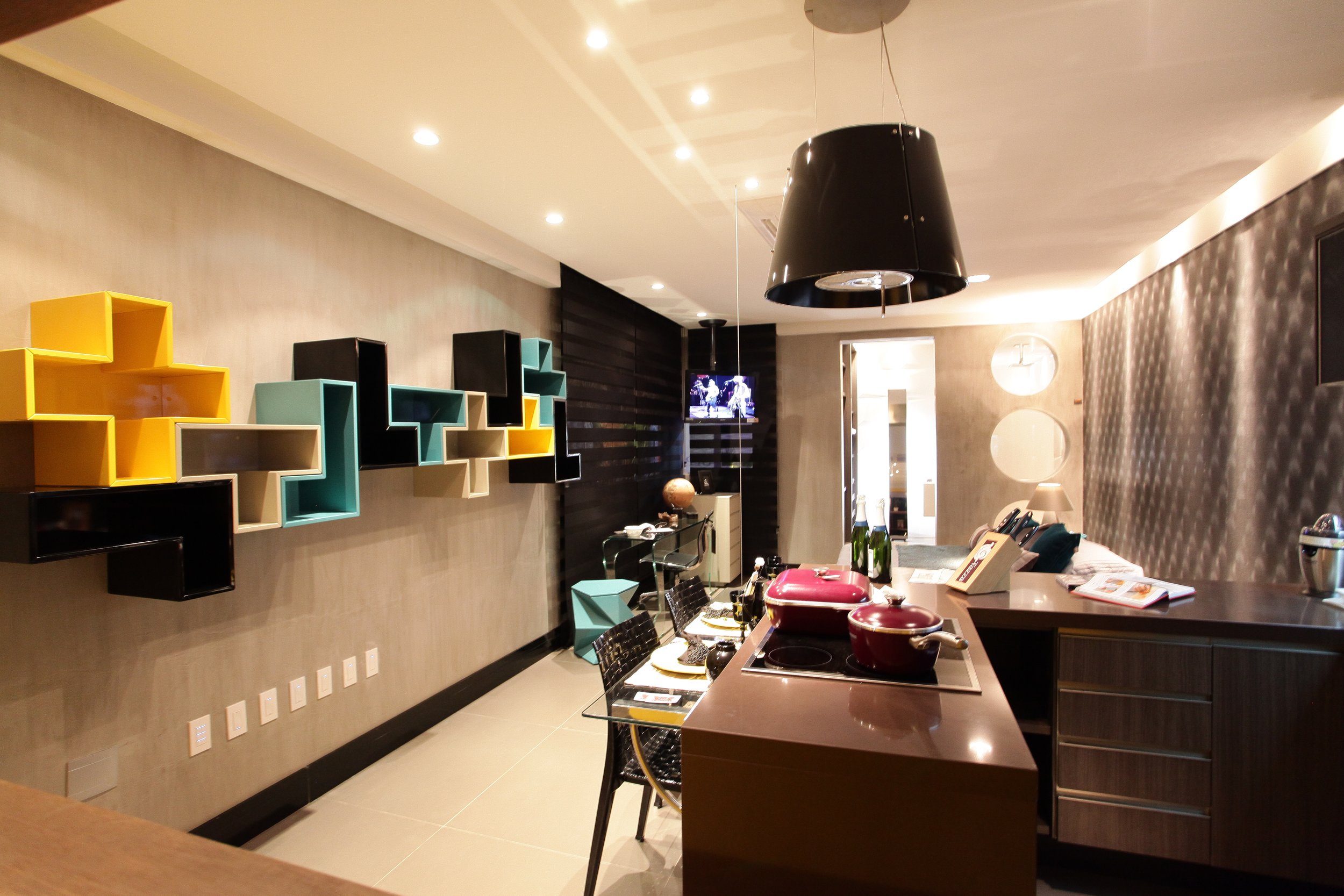 Modern kitchen with colorful geometric wall shelves, a black pendant lamp, a glass dining table with black chairs, and a kitchen counter with red cookware and bottles.