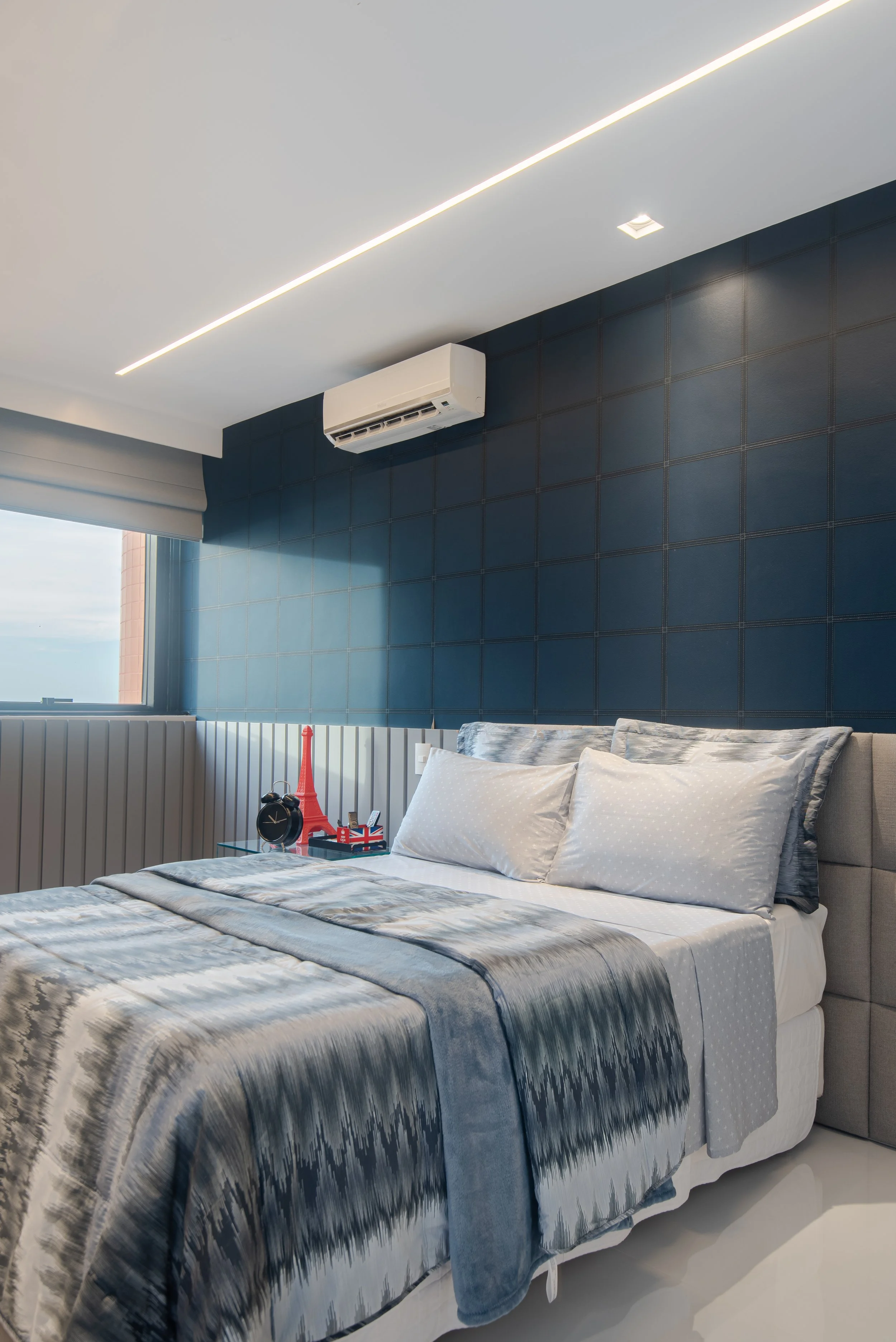 Modern bedroom with a large bed decorated with gray and white bedding, a bedside table with a small Eiffel Tower and Union Jack decoration, and a window with a view of a distant building. The room has a dark blue accent wall, a white ceiling, and a s