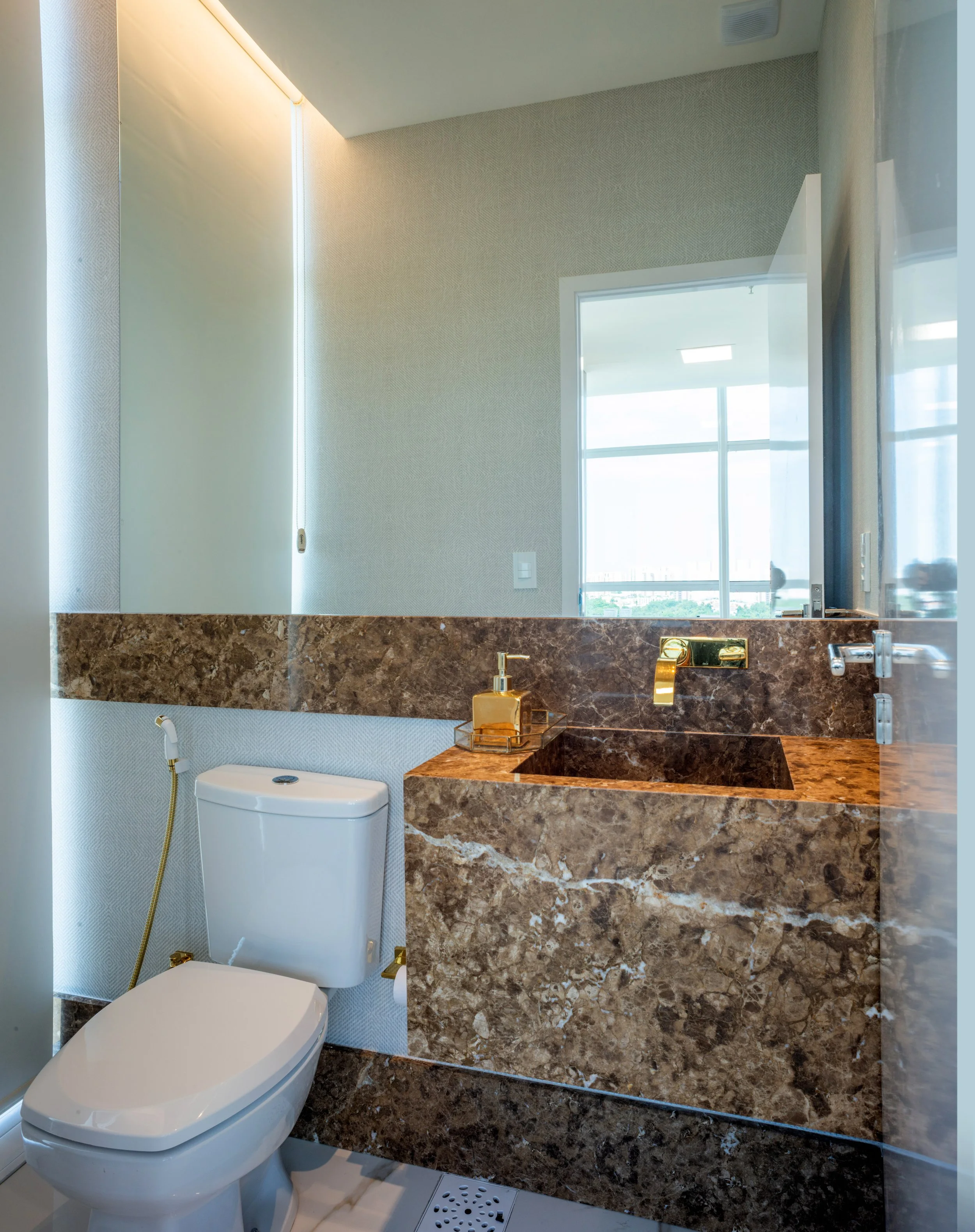 Modern bathroom with marble vanity, gold fixtures, toilet, and a window with natural light.