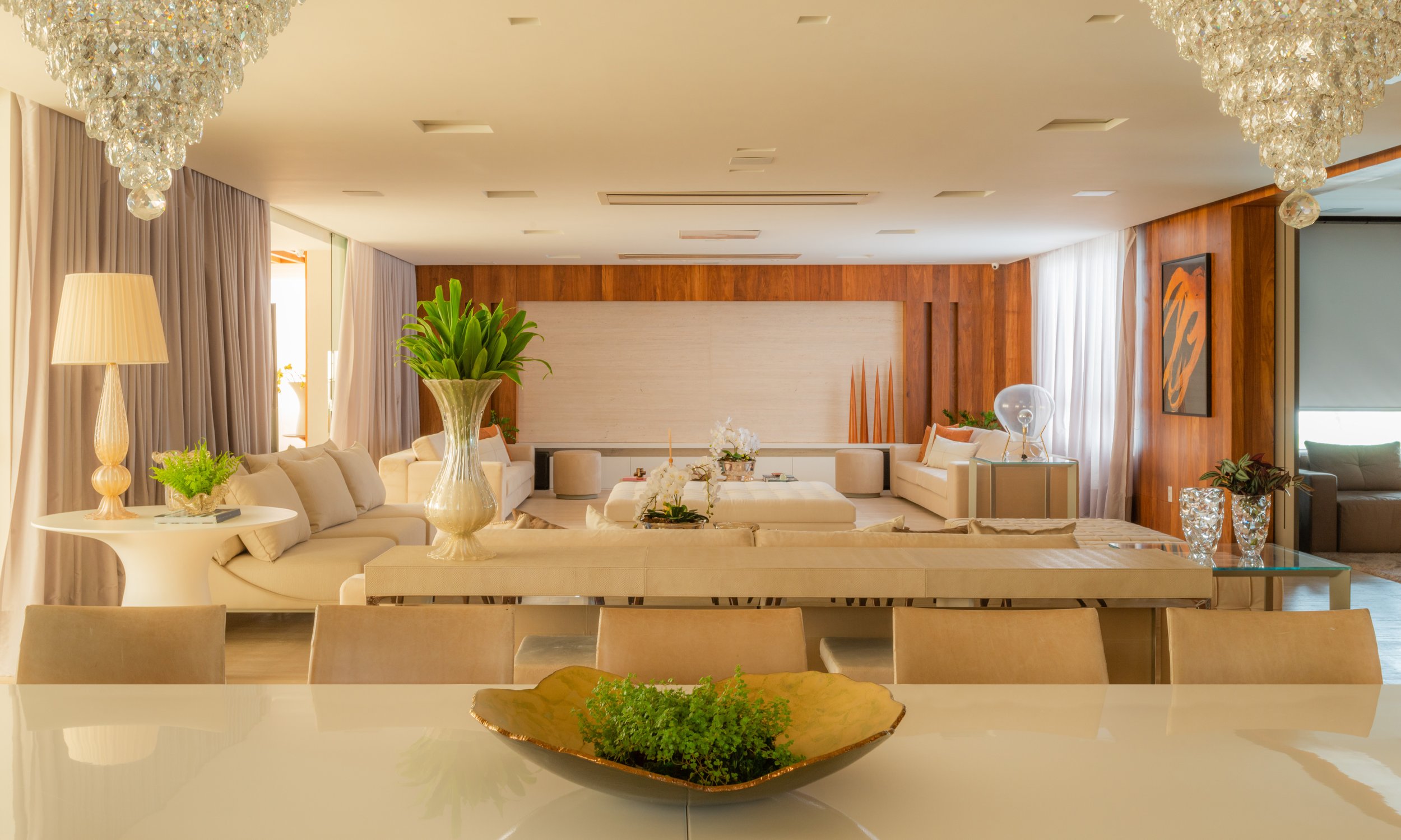 Elegant living room with white sofas, glass coffee table, green plants, decorative vases, wooden wall accents, and large windows with sheer curtains