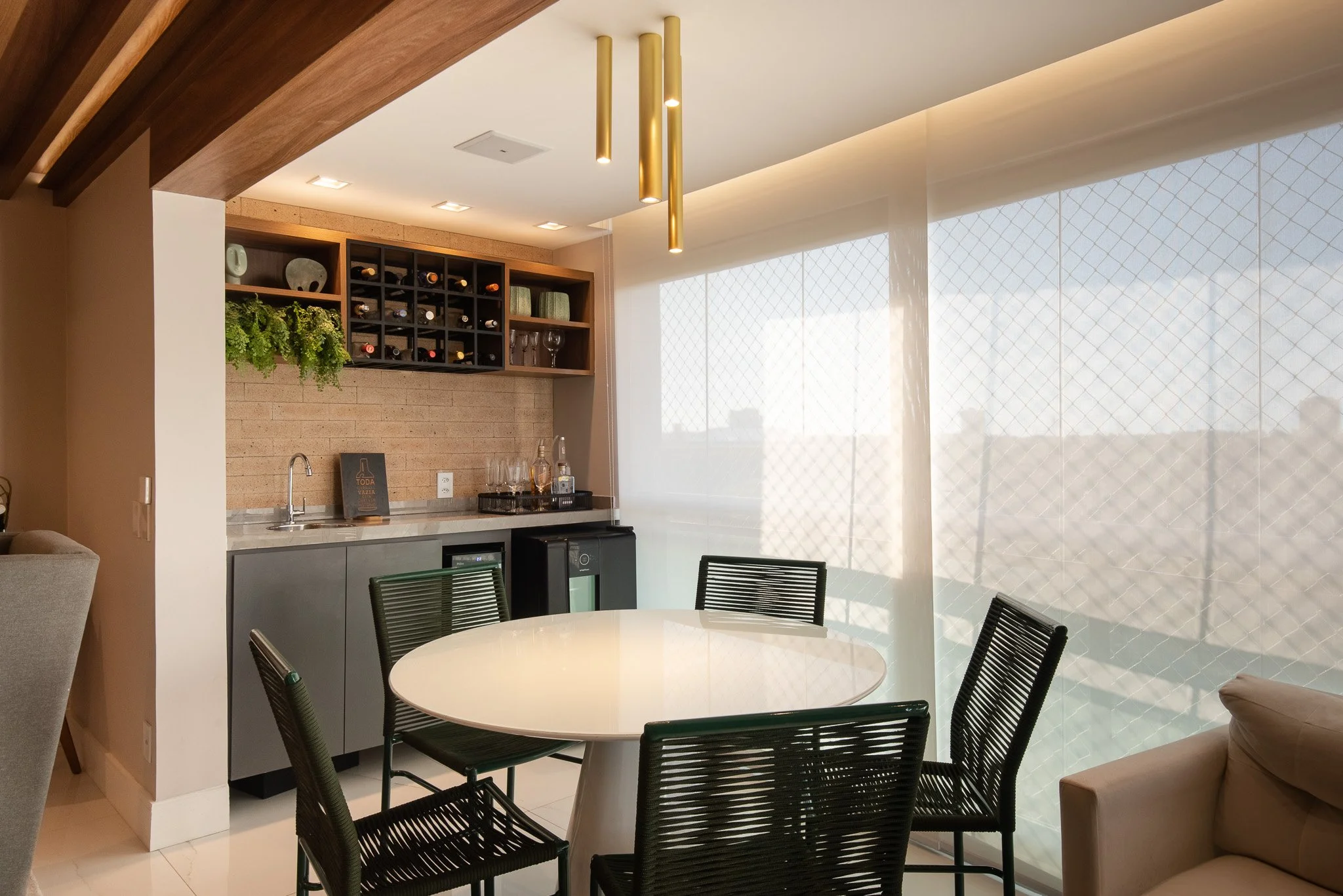 Modern dining area with a round white table and black chairs, with a small bar area and large windows covered with sheer curtains in the background.
