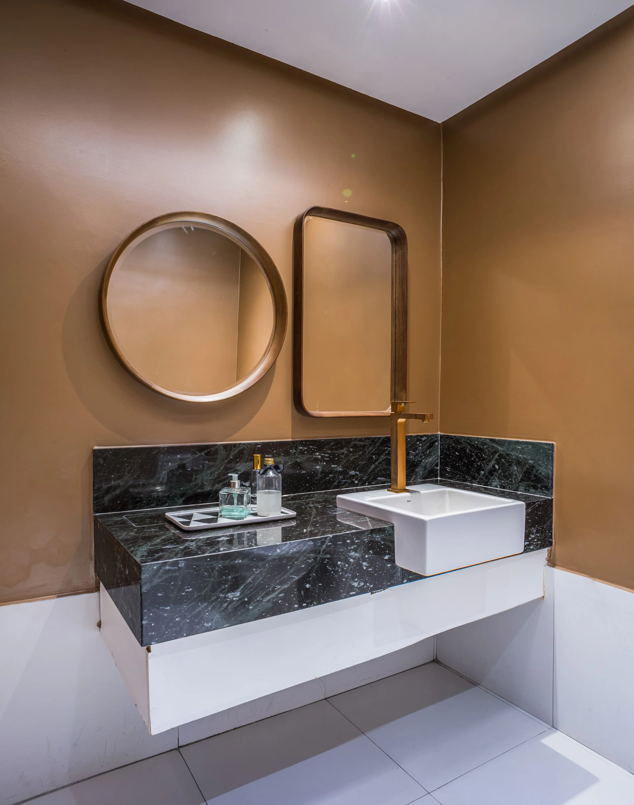 Modern bathroom sink with black marble countertop, a rectangular white vessel sink, and gold faucet, with round and rectangular mirrors on beige wall.