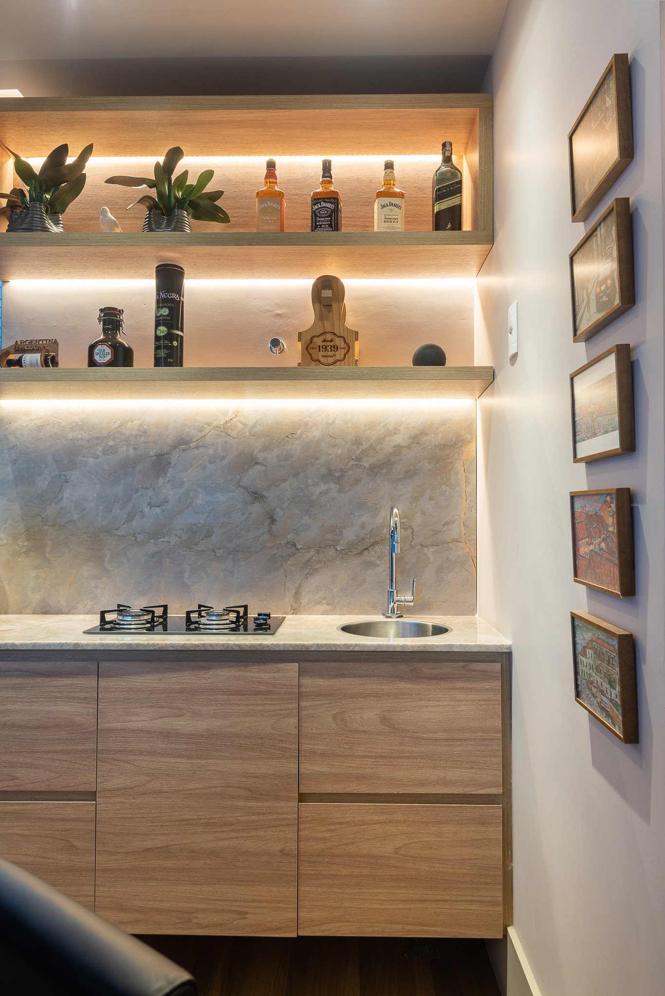 A modern kitchen with a marble backsplash, wooden cabinets, a small sink, a two-burner stove, and shelves above holding bottles, plants, and small decor items, with framed pictures on the right wall.