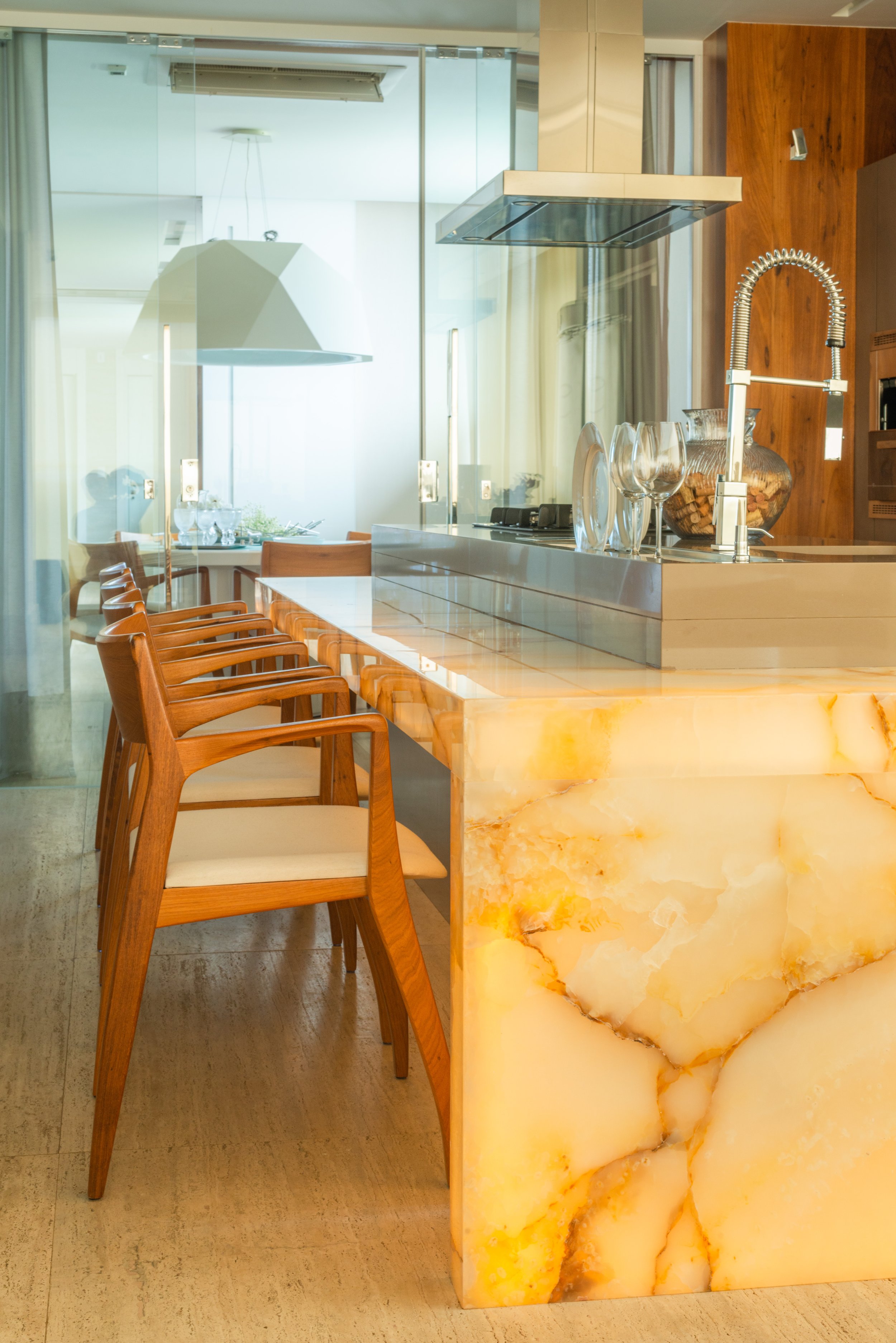 Modern kitchen with marble island, wooden chairs, glass wall, and a visible dining area in the background.