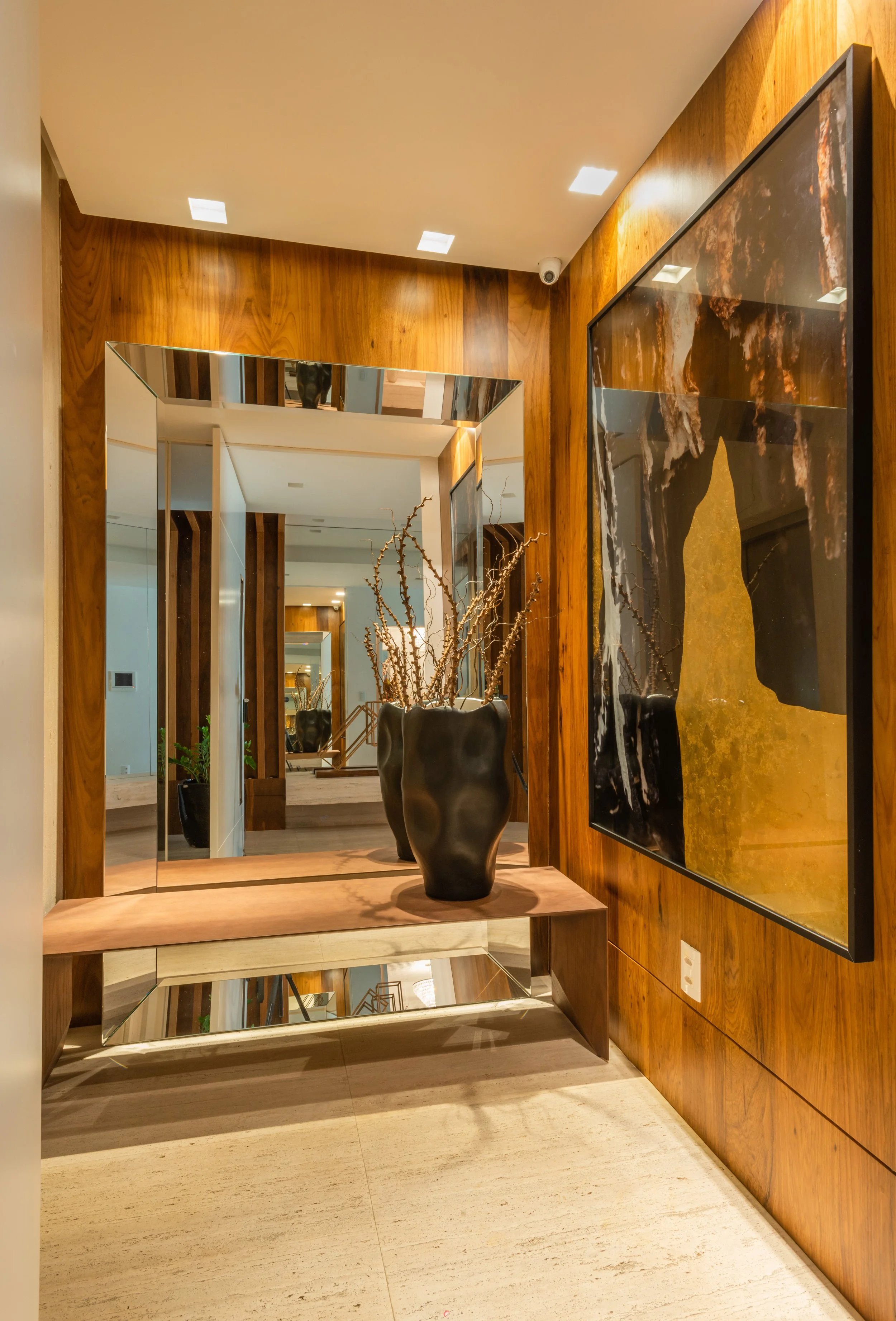 Modern interior hallway with wooden paneled walls, a large mirror, a decorative black vase with dried branches, a large painting on the wall, and a tall potted plant in the background.