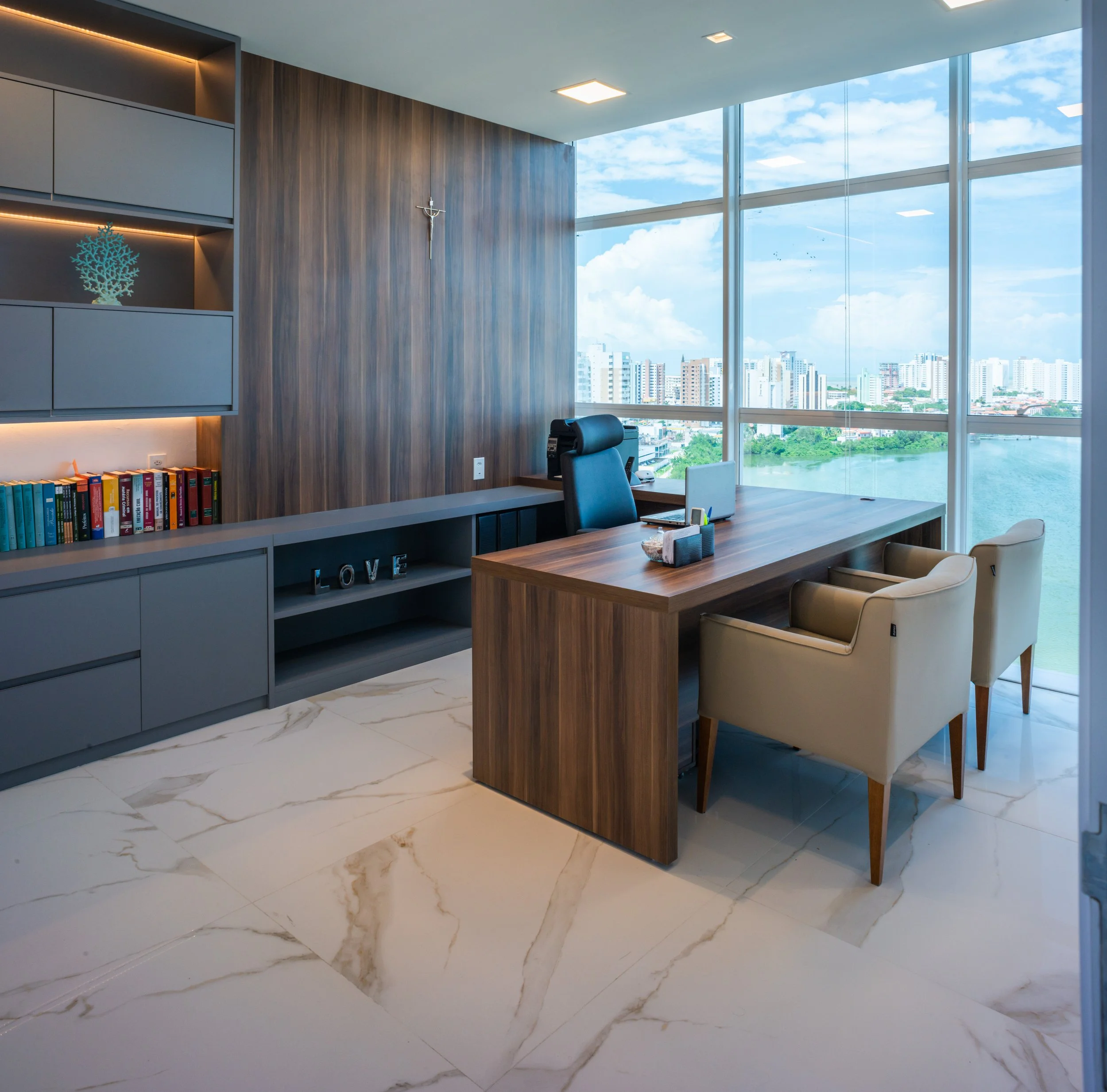 Modern office with wooden desk, beige chairs, large windows showing cityscape and water, books, decorative items, and a crucifix on the wall.