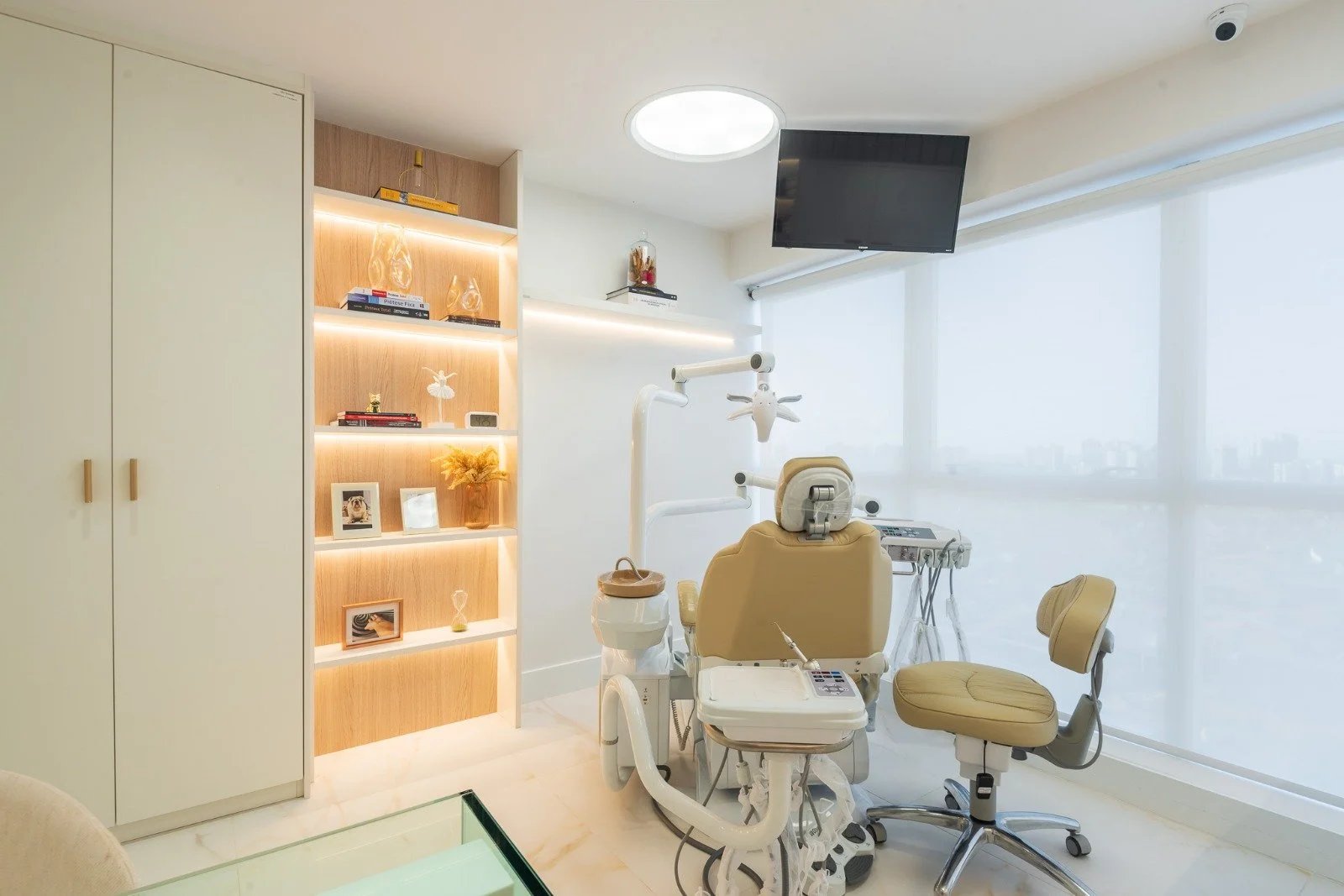 Dental examination chair in a bright, modern dental office, with a TV mounted on the ceiling and a large window with white blinds.