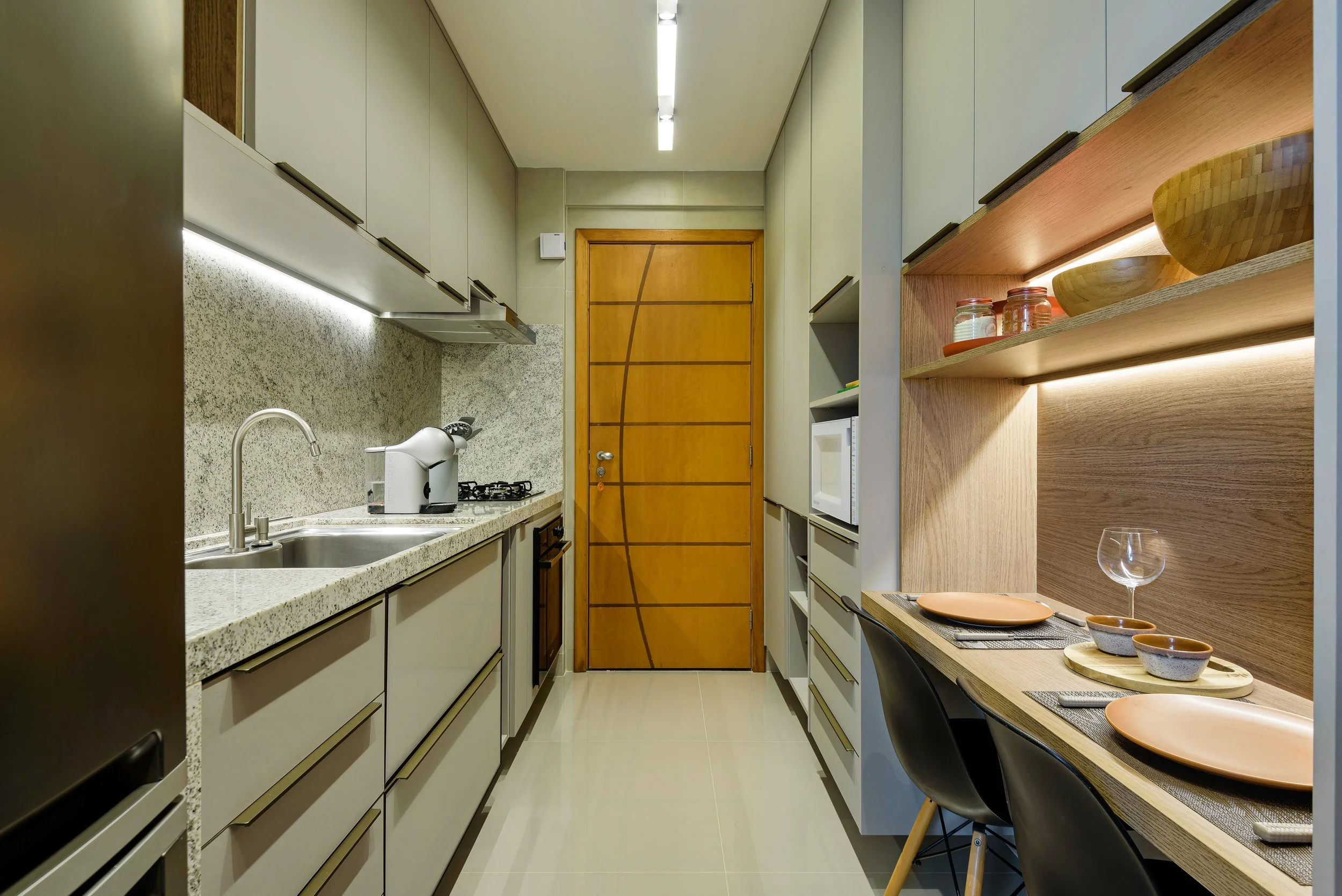 Modern galley kitchen with beige cabinetry, granite countertops, and a wooden door at the end. Includes a sink, microwave, stove, and tabletop dining area with plates, bowls, and wine glass.