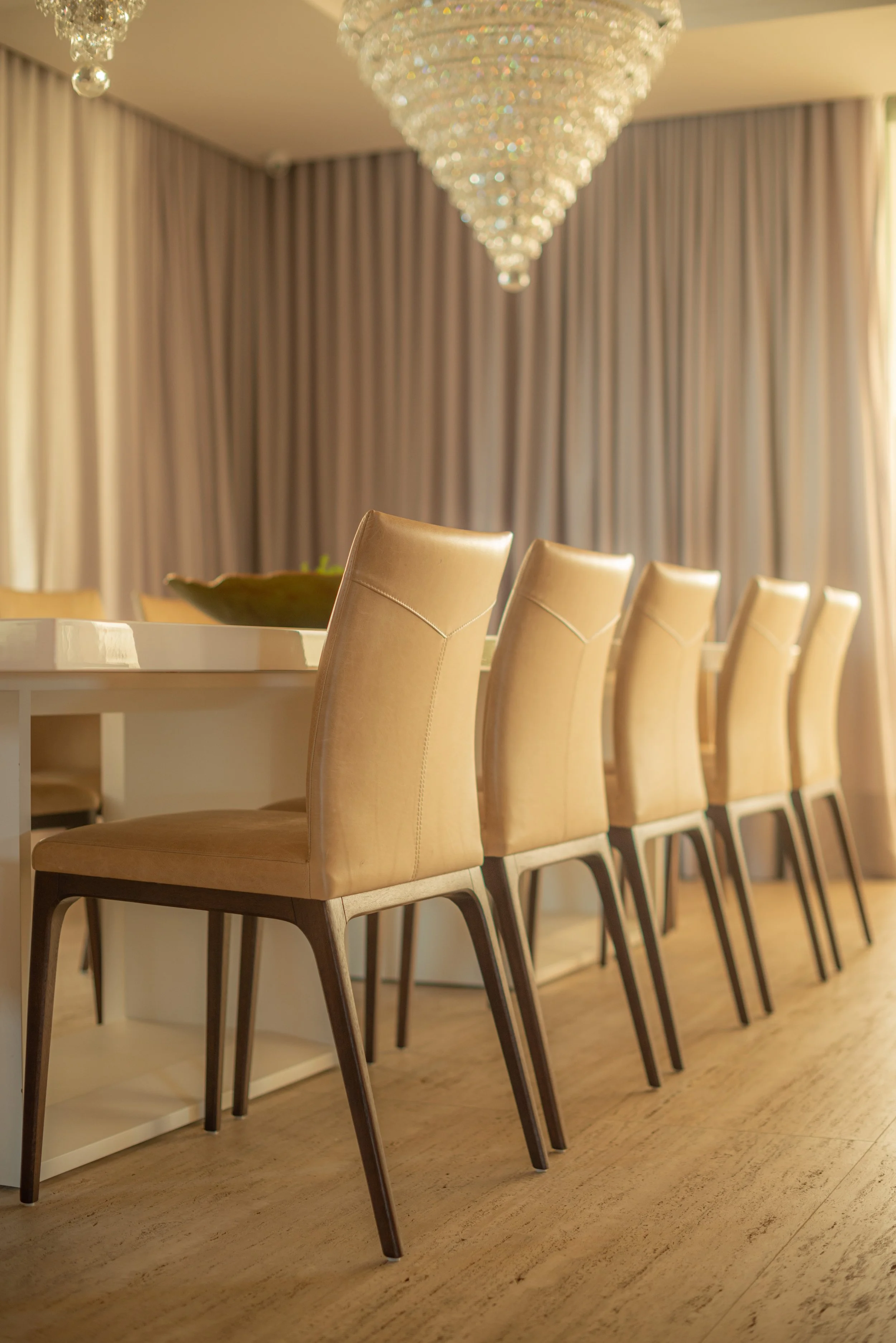 A row of six beige dining chairs with dark wooden legs positioned along one side of a white dining table in a well-lit room with beige curtains and a large, elegant chandelier hanging from the ceiling.