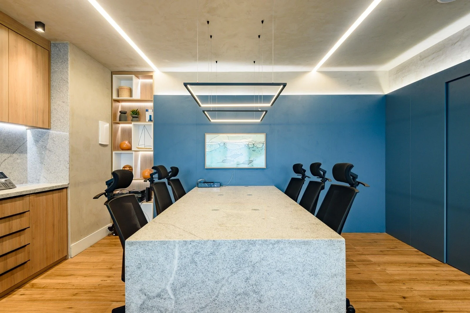 Modern conference room with a large granite table, black ergonomic chairs, blue accent wall with a framed aquatic-themed painting, wood accents, built-in shelves with decorative items, and contemporary lighting fixtures.