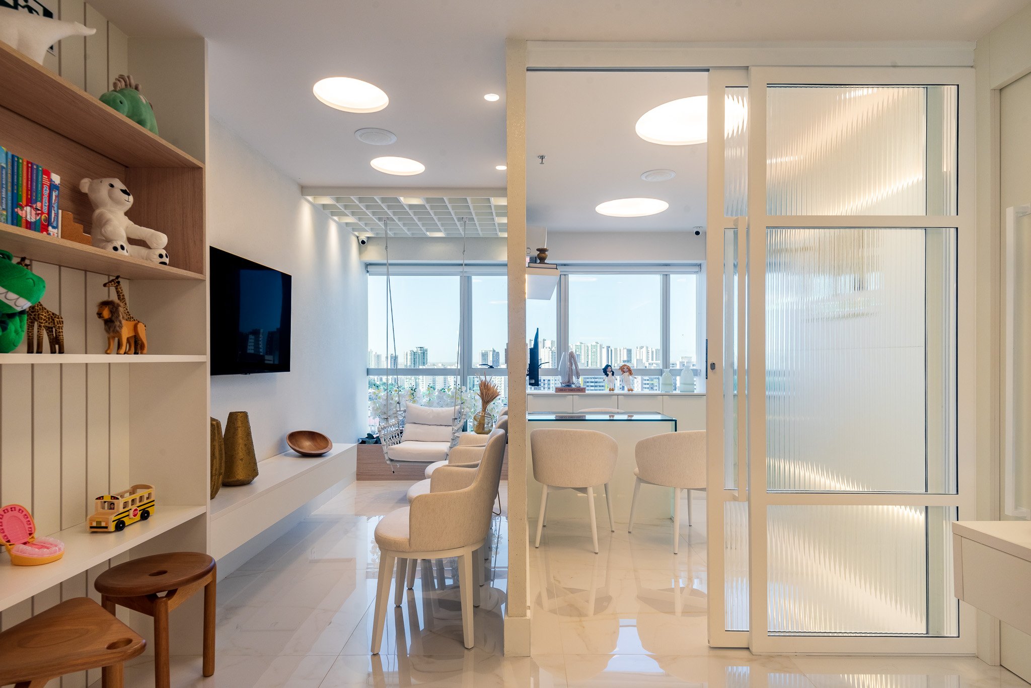 Bright, modern living space with white walls and furniture, large windows providing city views, a shelf with children's toys, a flat-screen TV, and a dining area with four cream-colored chairs.