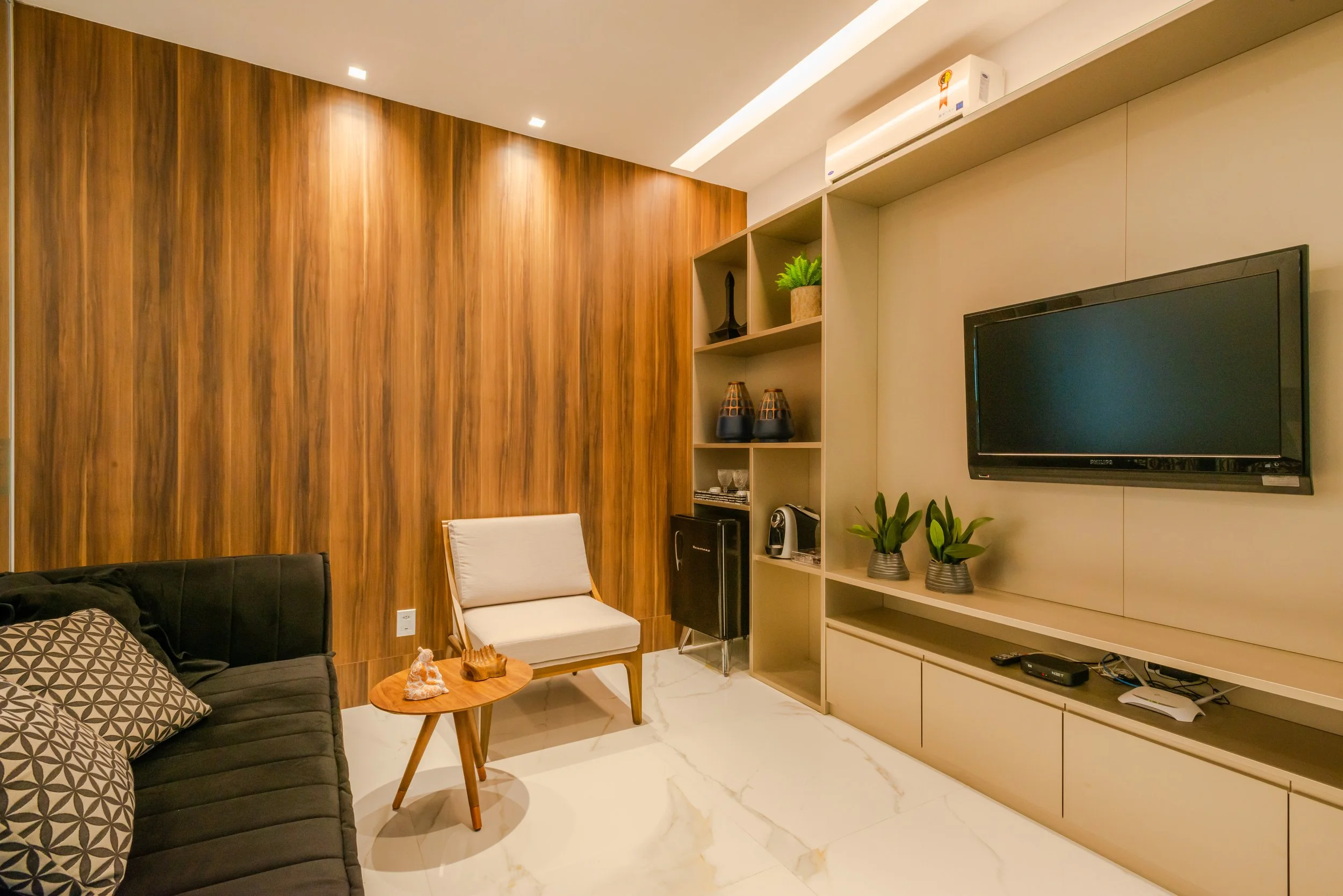 Living room with a black sofa, a wooden side table, and a white armchair. There is a wall-mounted TV, potted plants, and decorative items on a beige entertainment unit. The wall behind the furniture has a wooden panel, and the floor is white marble.