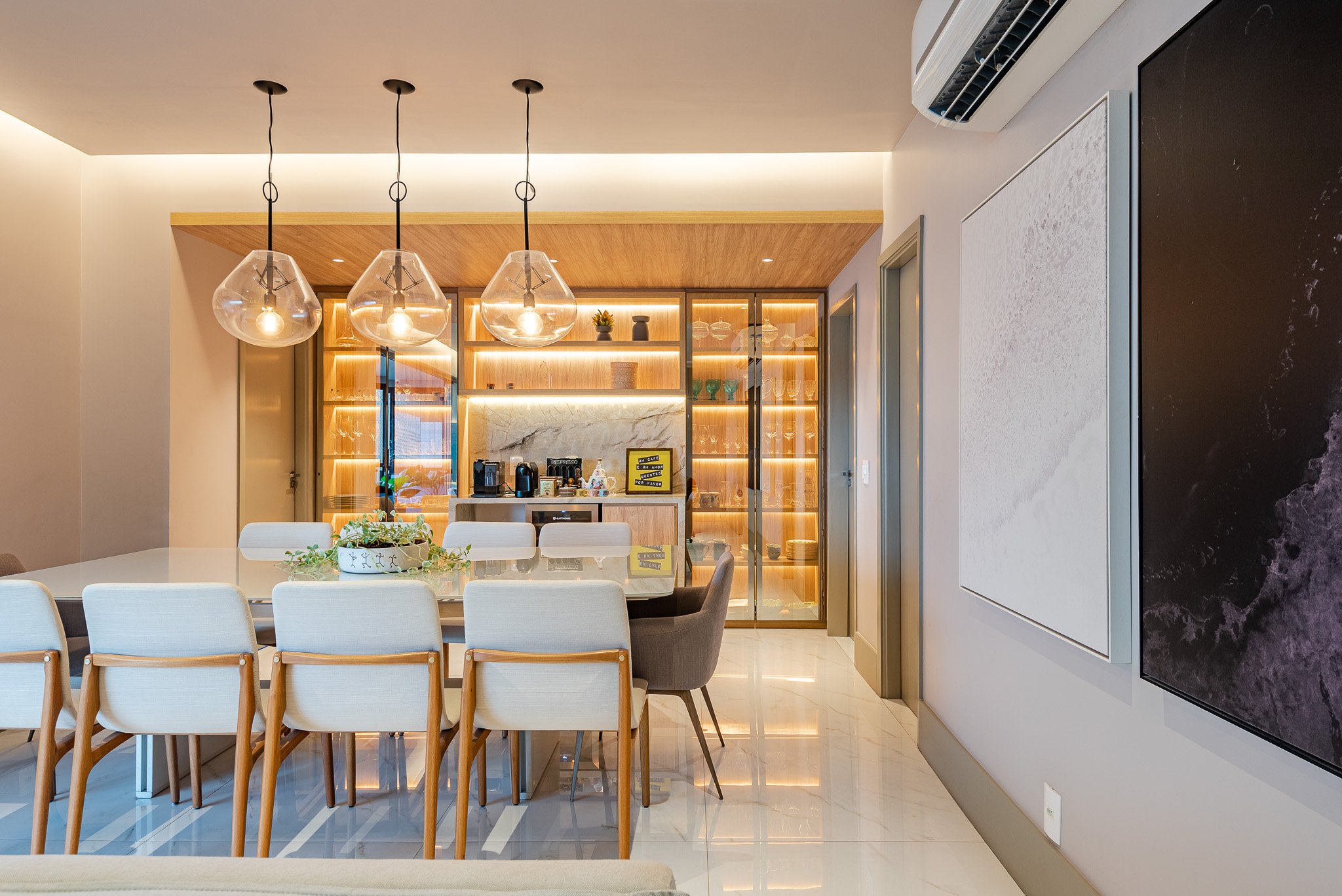 Modern dining area with a large glass-topped table, white chairs, and contemporary hanging light fixtures. In the background, a wooden and marble bar area with built-in lighting and shelves for glassware and decor.