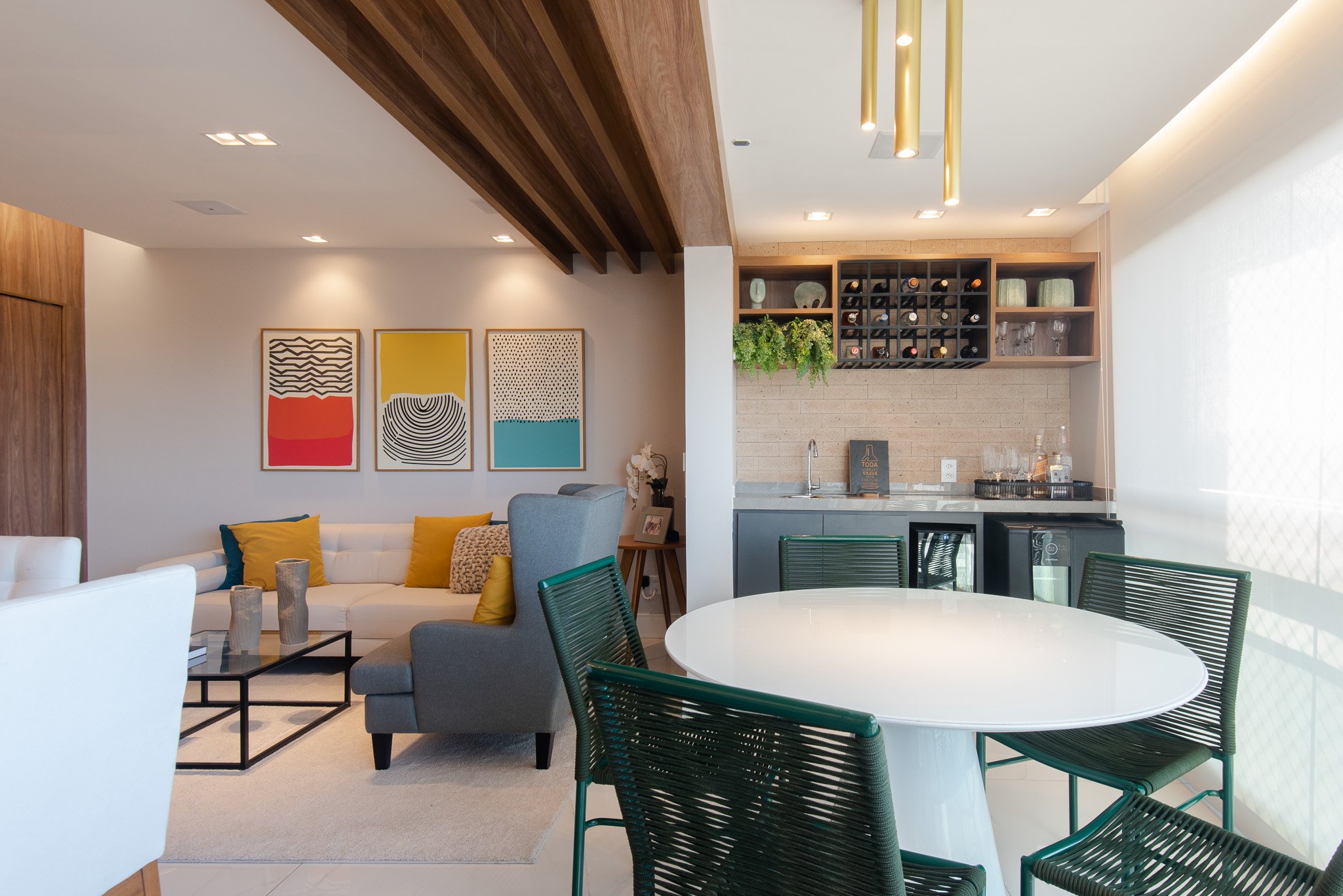 Open-concept living room and kitchen with modern decor, white walls, colorful artwork, yellow and gray pillows, a white sofa, green chairs around a white table, and a small kitchen area with wine storage and plants.