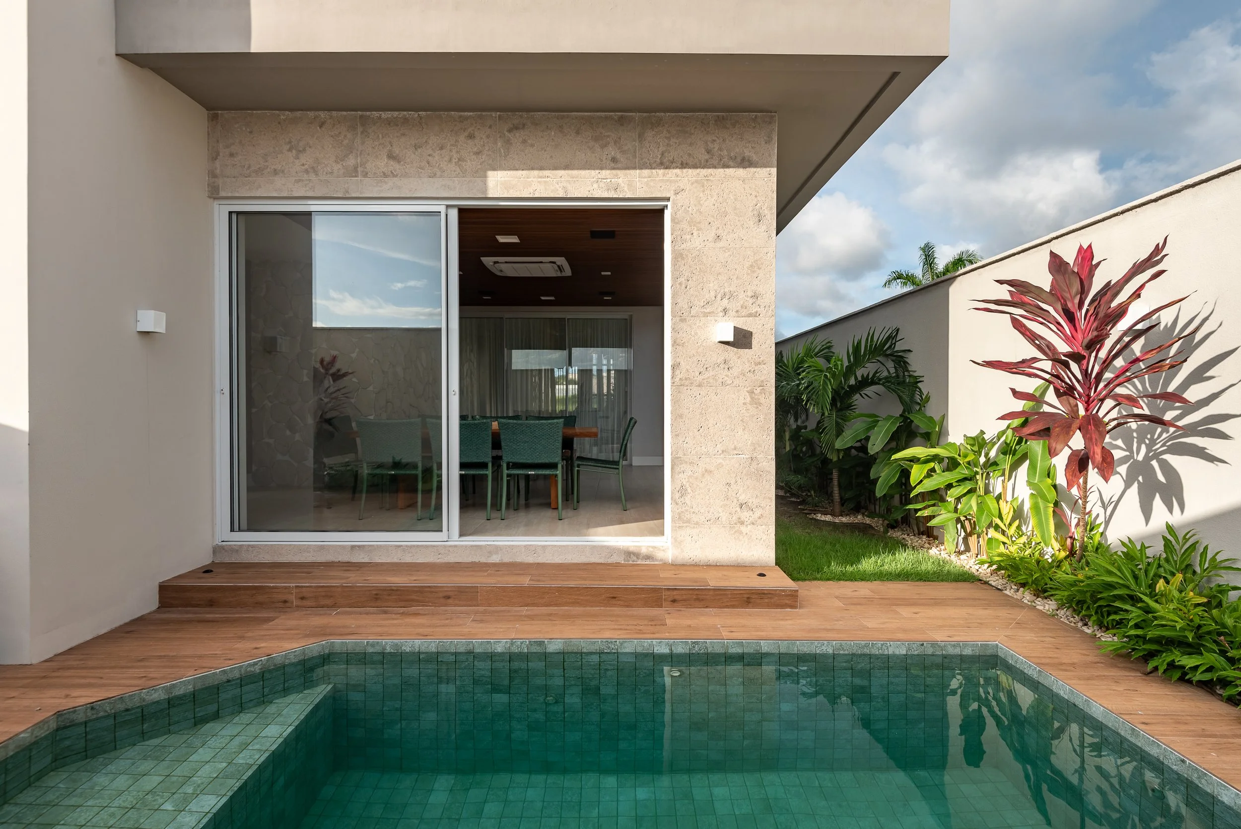 View of a modern backyard with a swimming pool, wooden deck, and patio door leading to an outdoor dining area inside a house.