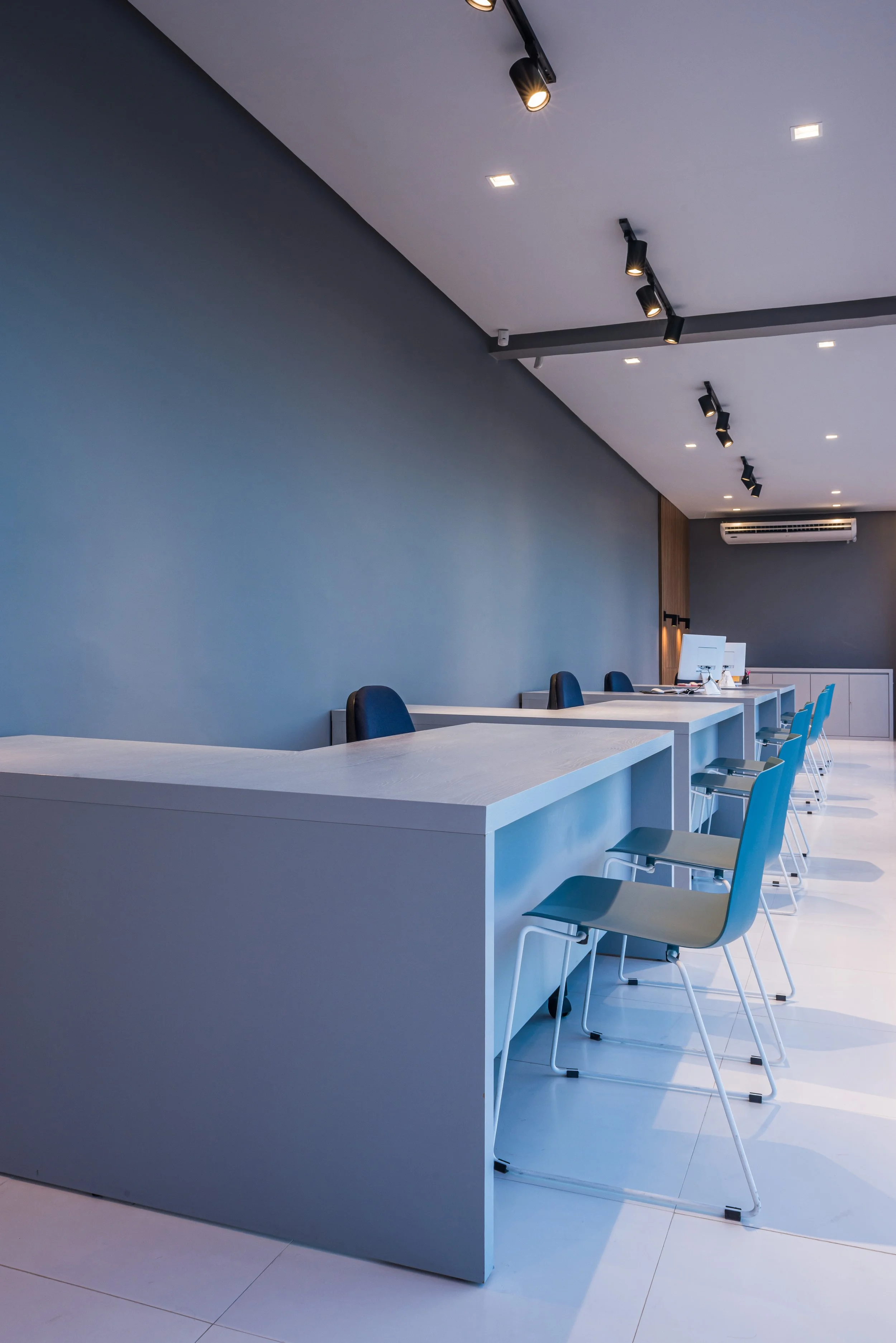 Modern reception or workspace with white counters, blue chairs, a computer, and track lighting on a gray wall ceiling.