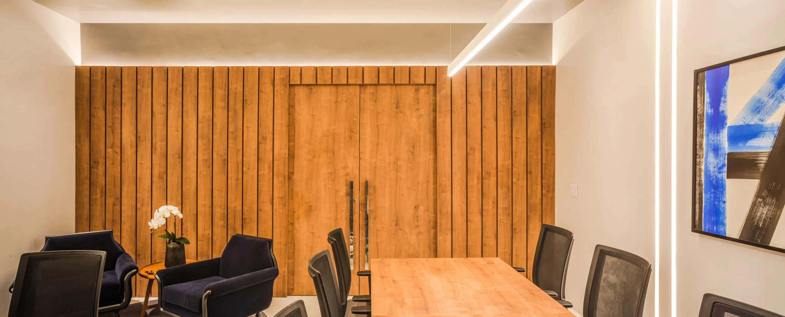 Modern conference room with wooden accents, a large table, black office chairs, a small side table with a orchid plant, and abstract artwork on the wall.