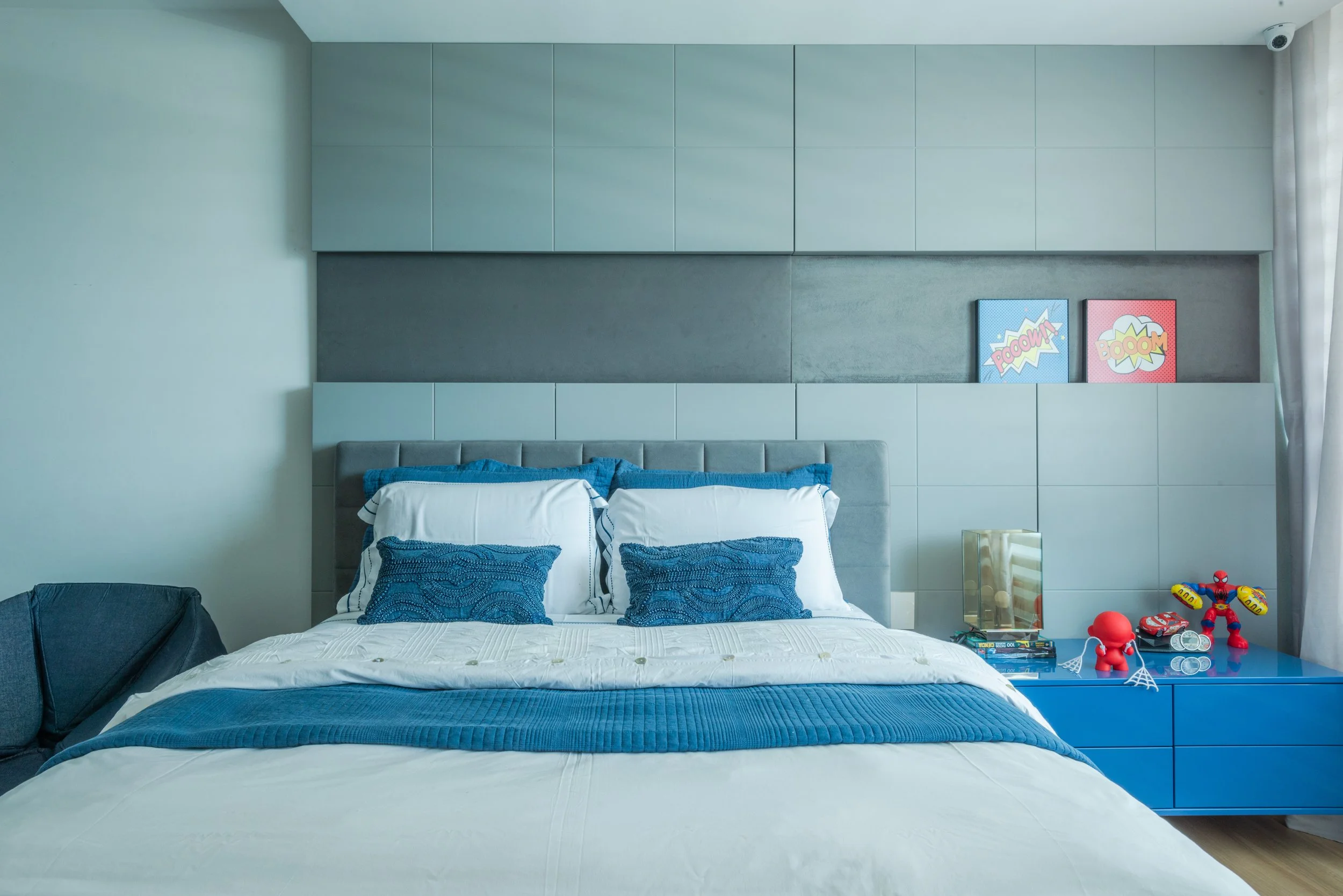 Modern bedroom with a bed, blue throw blanket, and decorative blue pillows. Blue dresser with toys, books, and comic art on the wall.