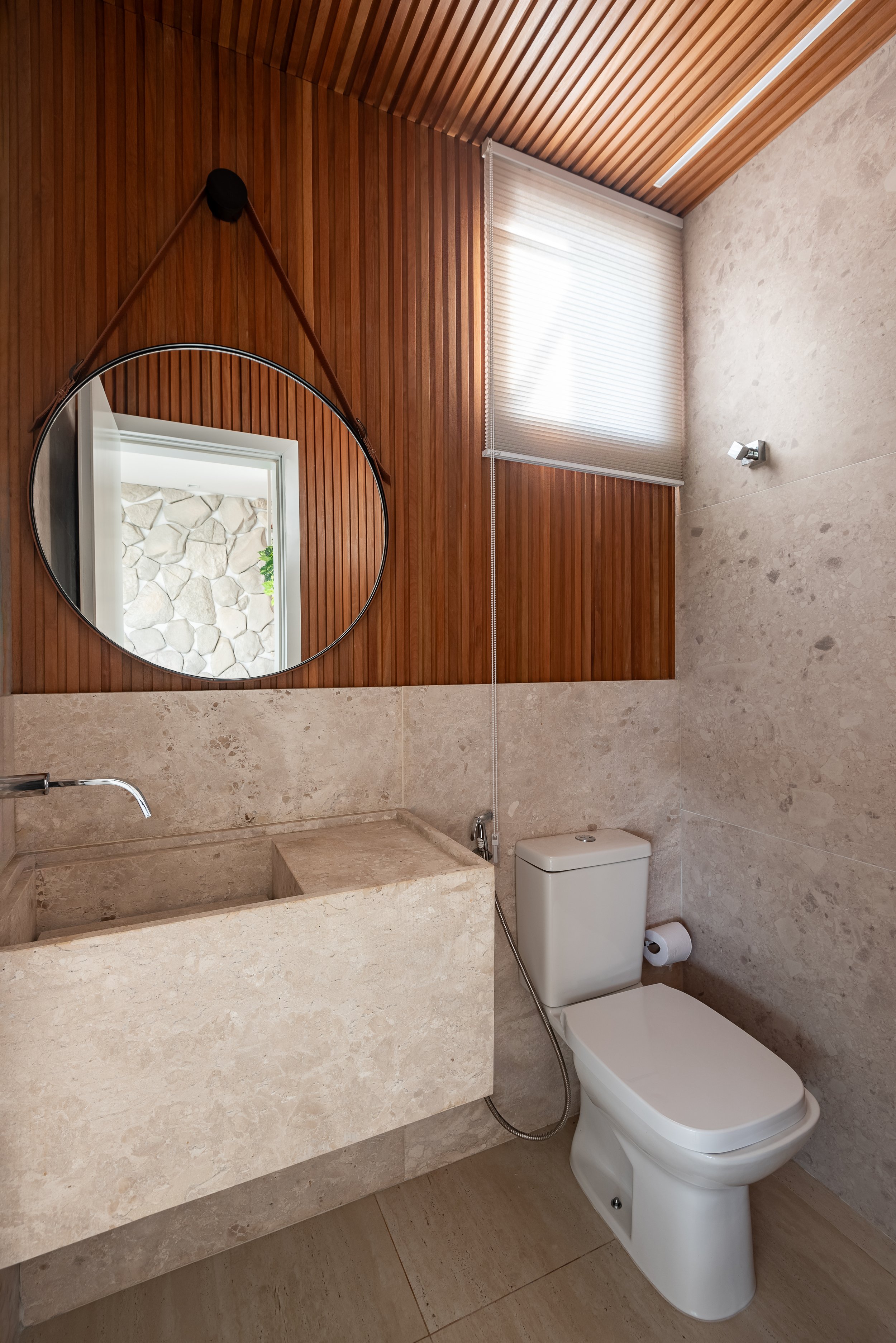 A modern bathroom with beige marble walls and countertop, a white toilet, a round mirror, and wood paneling on the upper wall and ceiling, with a window covered by a white blind.