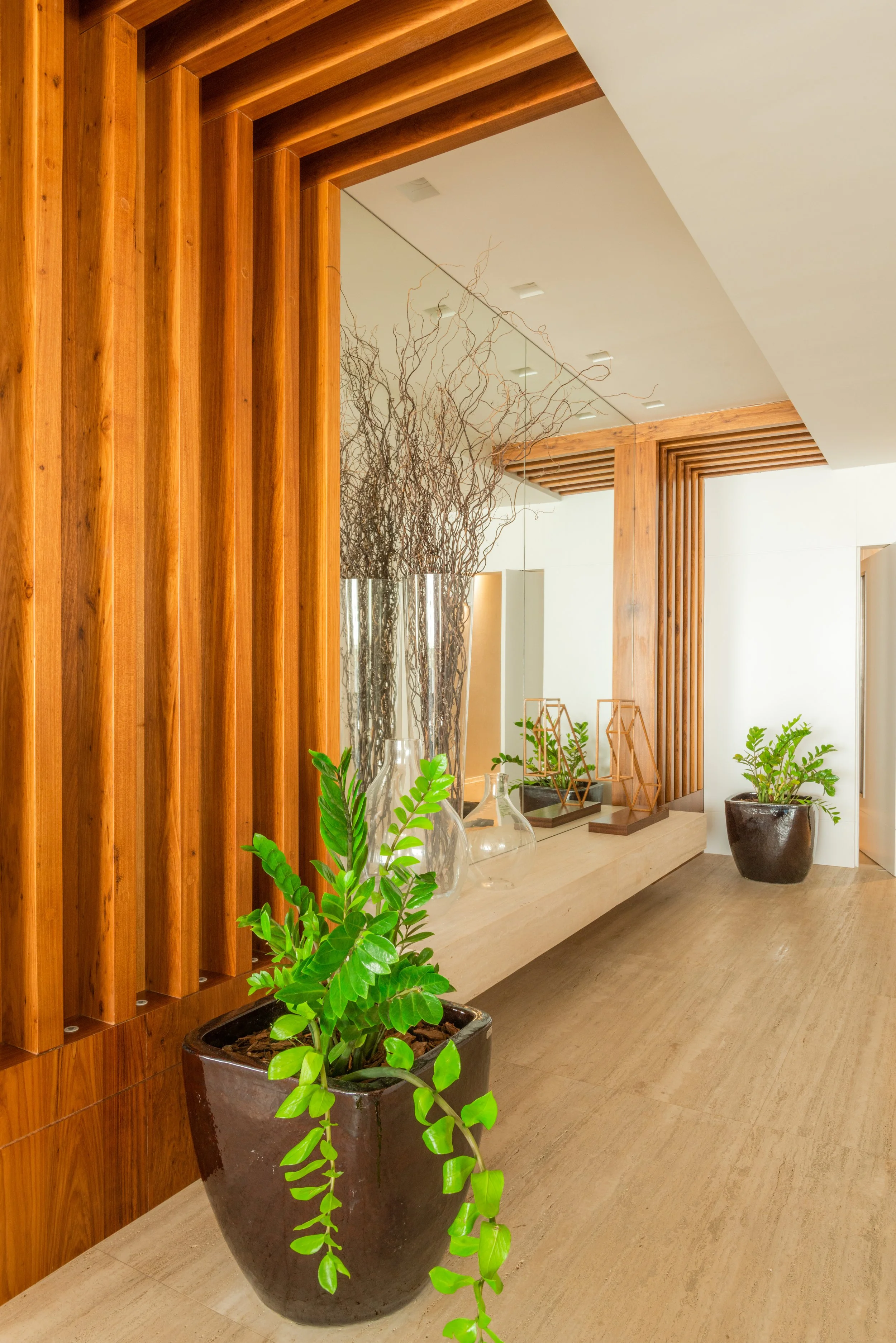 Decorative interior with large potted green plants, wooden wall panels, and a cream-colored bench with glass vases and geometric decor.