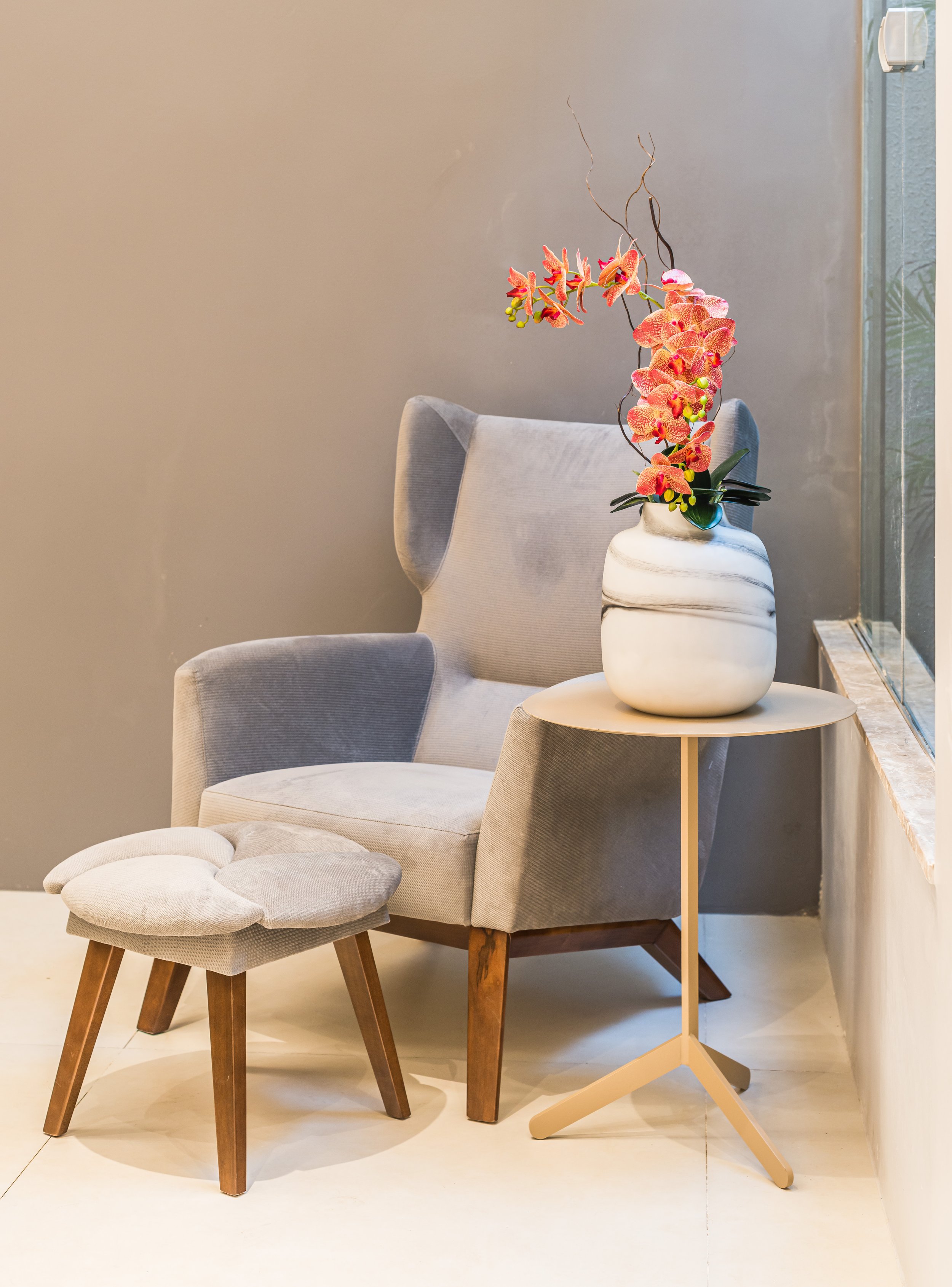 A cozy reading corner with a light gray wingback armchair, a matching ottoman, next to a small round beige side table with a white marble vase filled with pink and orange orchids, and a window on the right side.