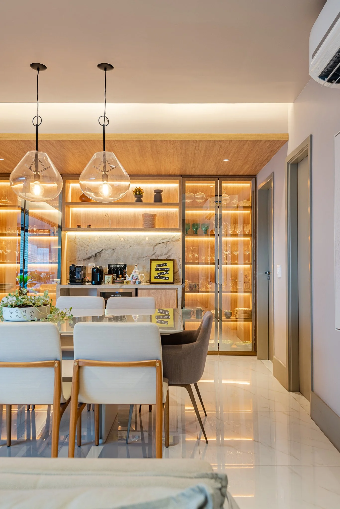 Modern dining area with a round table, six chairs, and a built-in bar with glass shelves illuminated with warm lighting in a contemporary home.