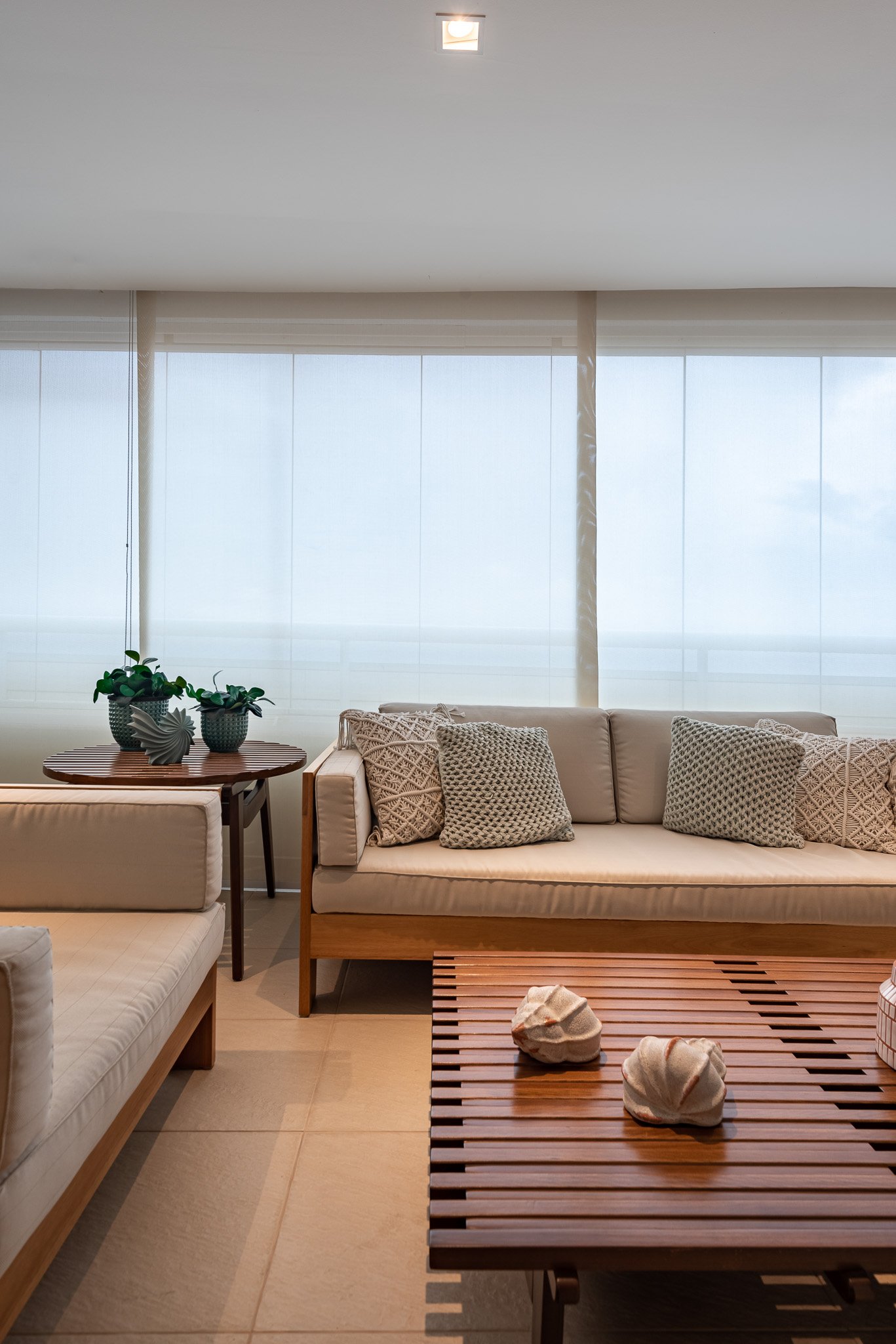 Living room with a white sofa and beige cushions, a wooden coffee table with shells on it, and a windows with sheer curtains.