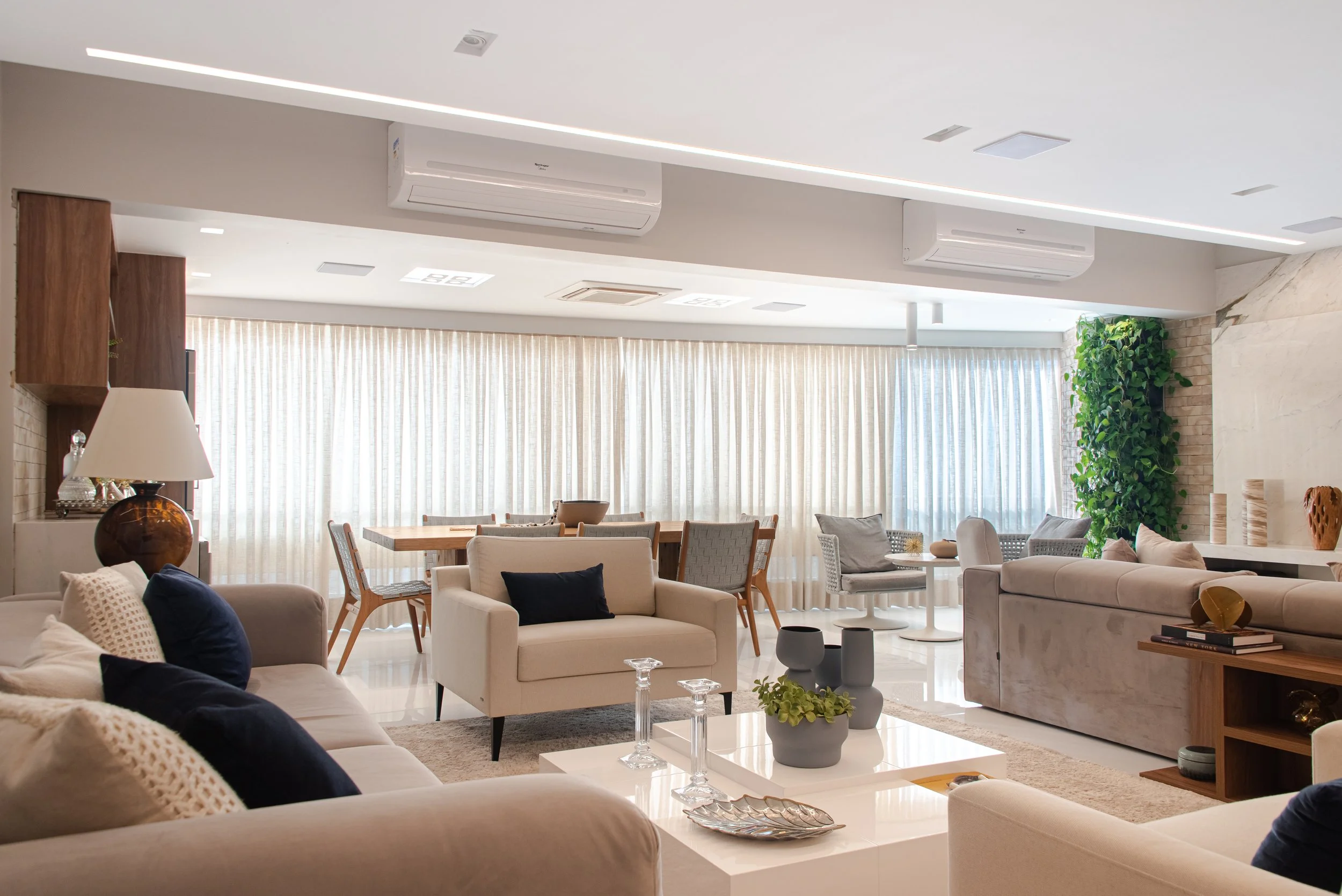 A spacious, modern living room with beige sofas, decorative pillows, a coffee table with vases, and a dining area with a wooden table and chairs. Large windows with light curtains and a green plant in the corner.