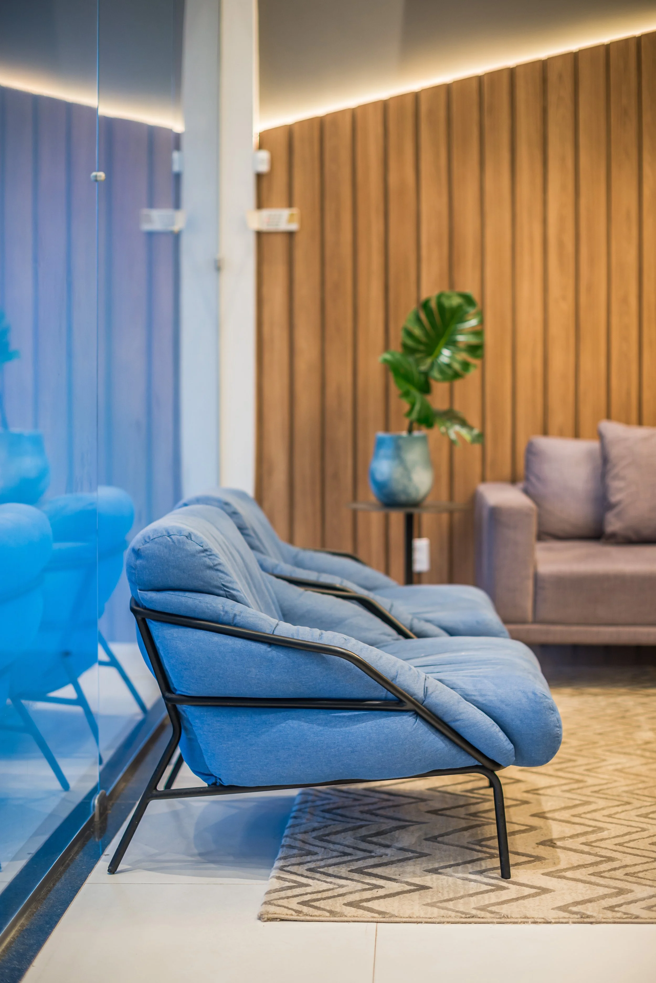 Modern waiting area with blue cushioned chairs, beige sofa, wooden wall, and potted plant.