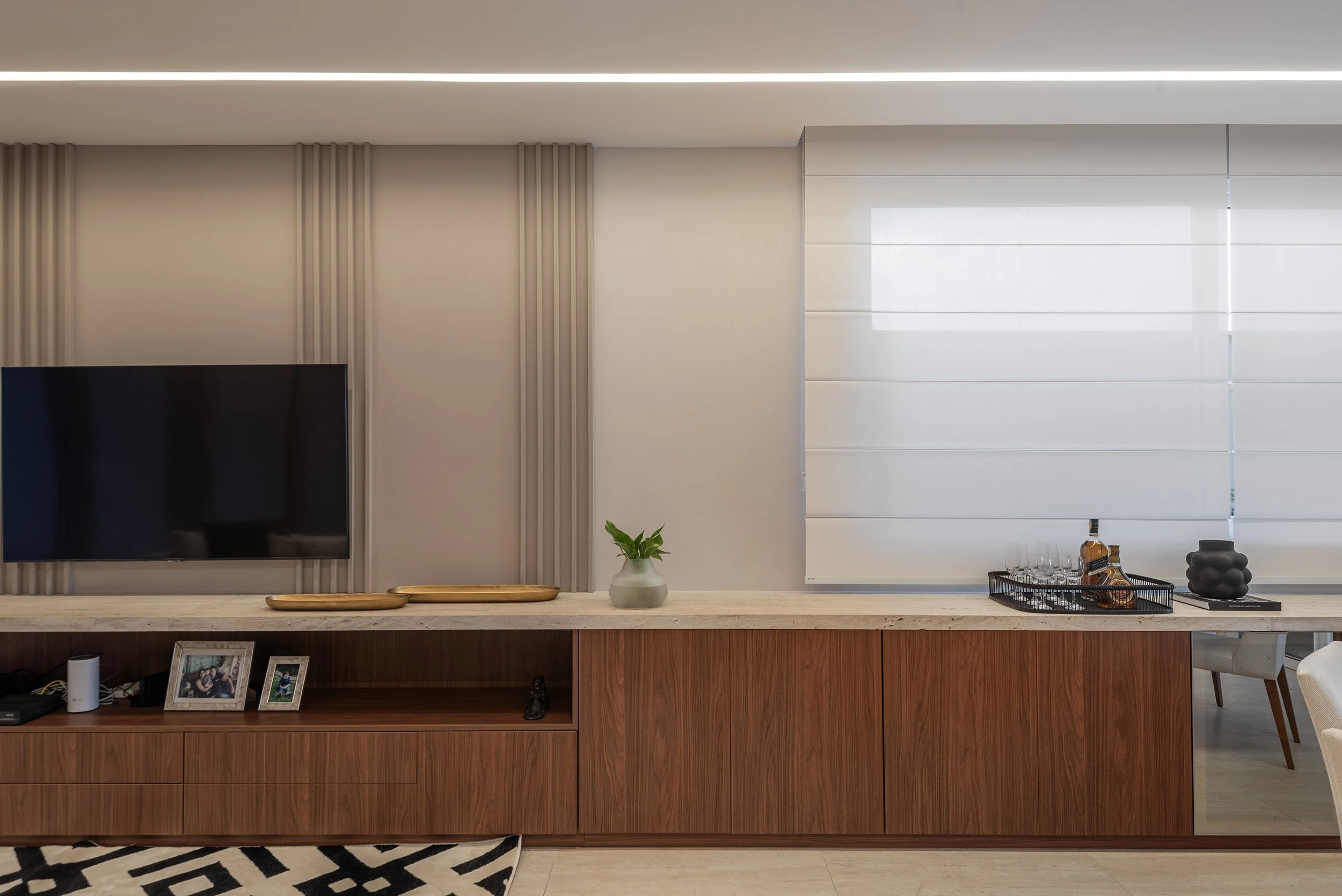 Modern living room with a wall-mounted flat-screen TV, a wooden console with pictures and decor, a small plant, and a window with white blinds.