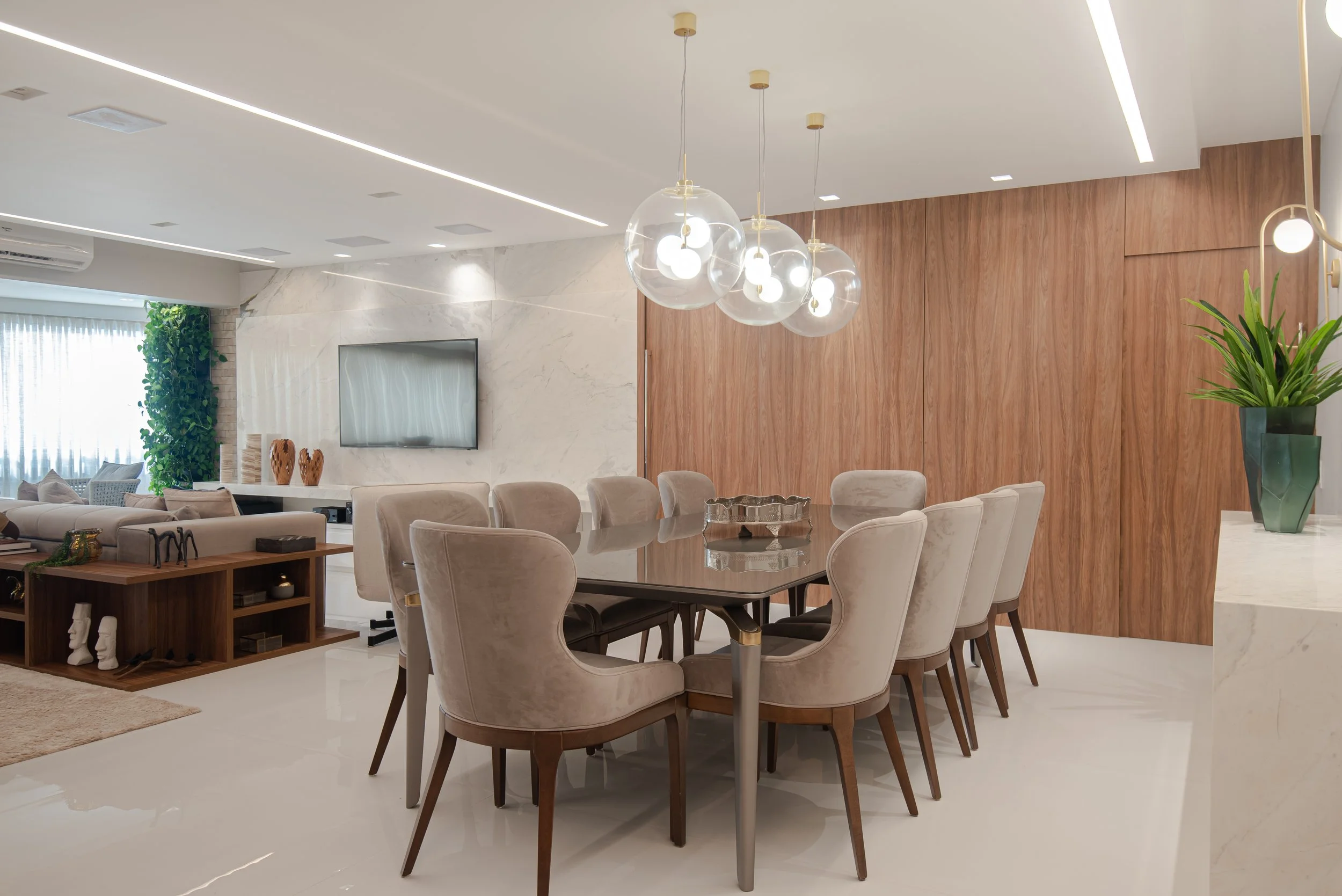 Modern dining room with beige upholstered chairs around a wooden table, a pendant light fixture with glass globes, and a wooden accent wall. The room features white walls, a marble wall with a TV, and a corner with a green plant.