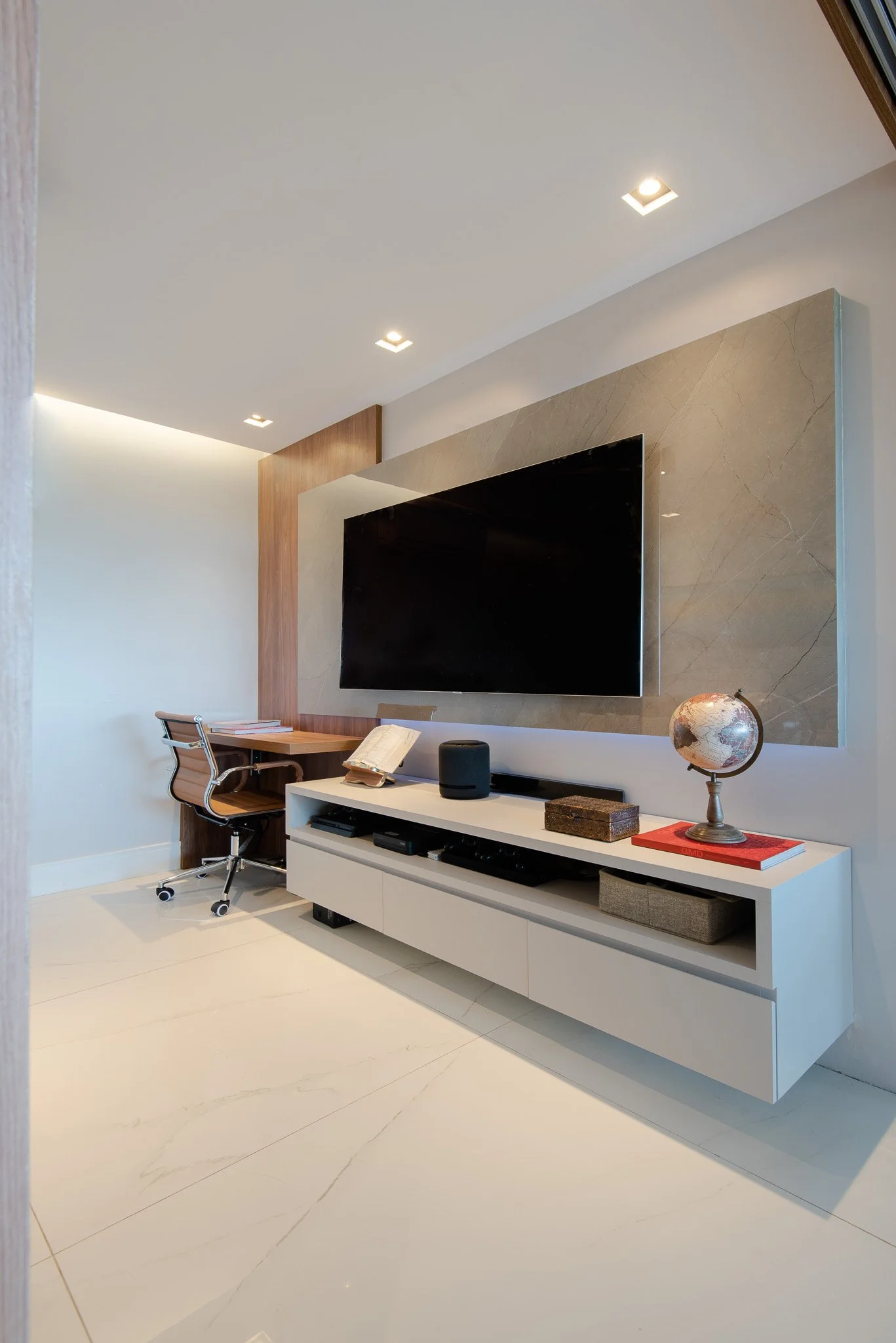 Modern living room with a wall-mounted flat-screen TV above a white entertainment console, a globe, books, and electronic devices, with a small wooden desk and chair in the corner.