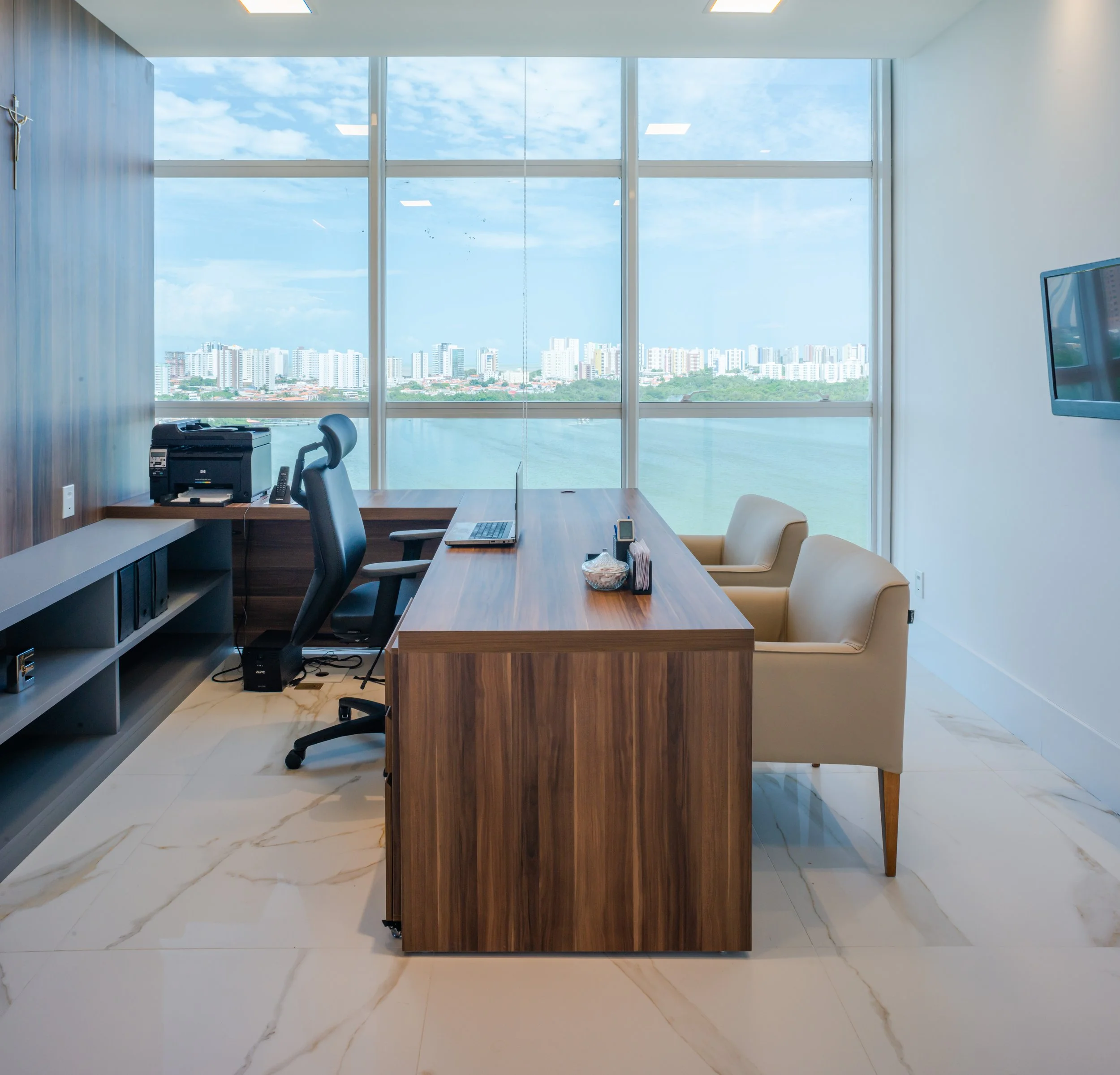 Modern office with large window overlooking city skyline and water, wooden desk with office chair, two beige armchairs, wall-mounted TV, printer, and various office supplies.